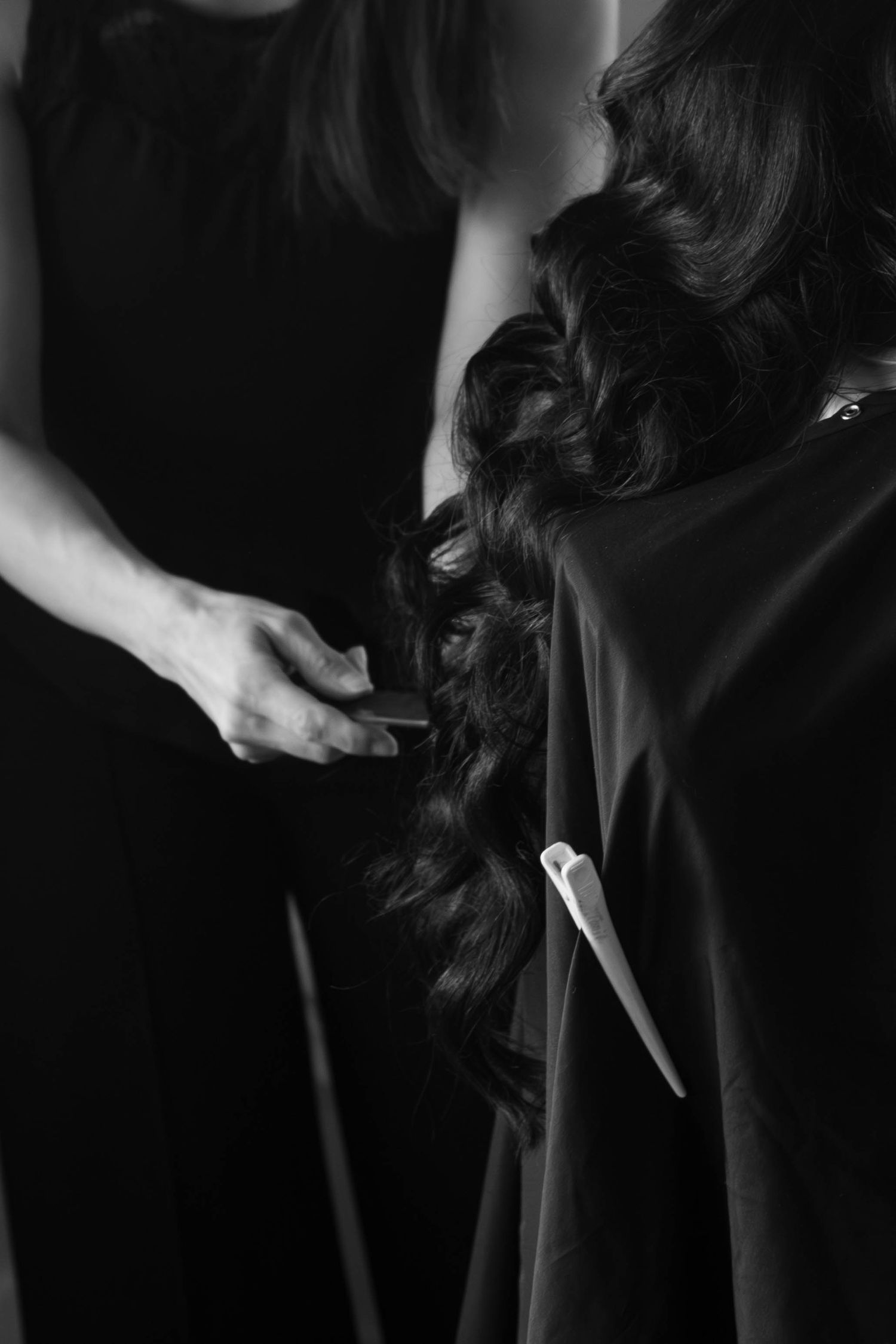 Black and white photo of a hairstylist styling long curly dark hair on a woman in a black dress.