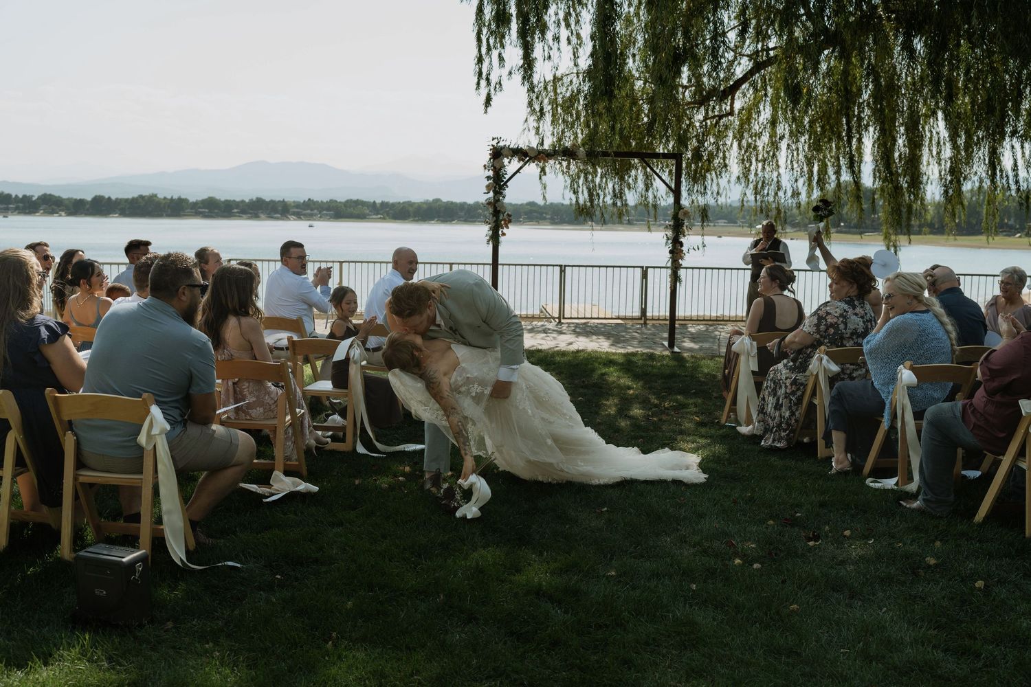 Outdoor wedding ceremony by a lake with bride and groom kissing at the altar, guests seated on chairs in lush green grass.