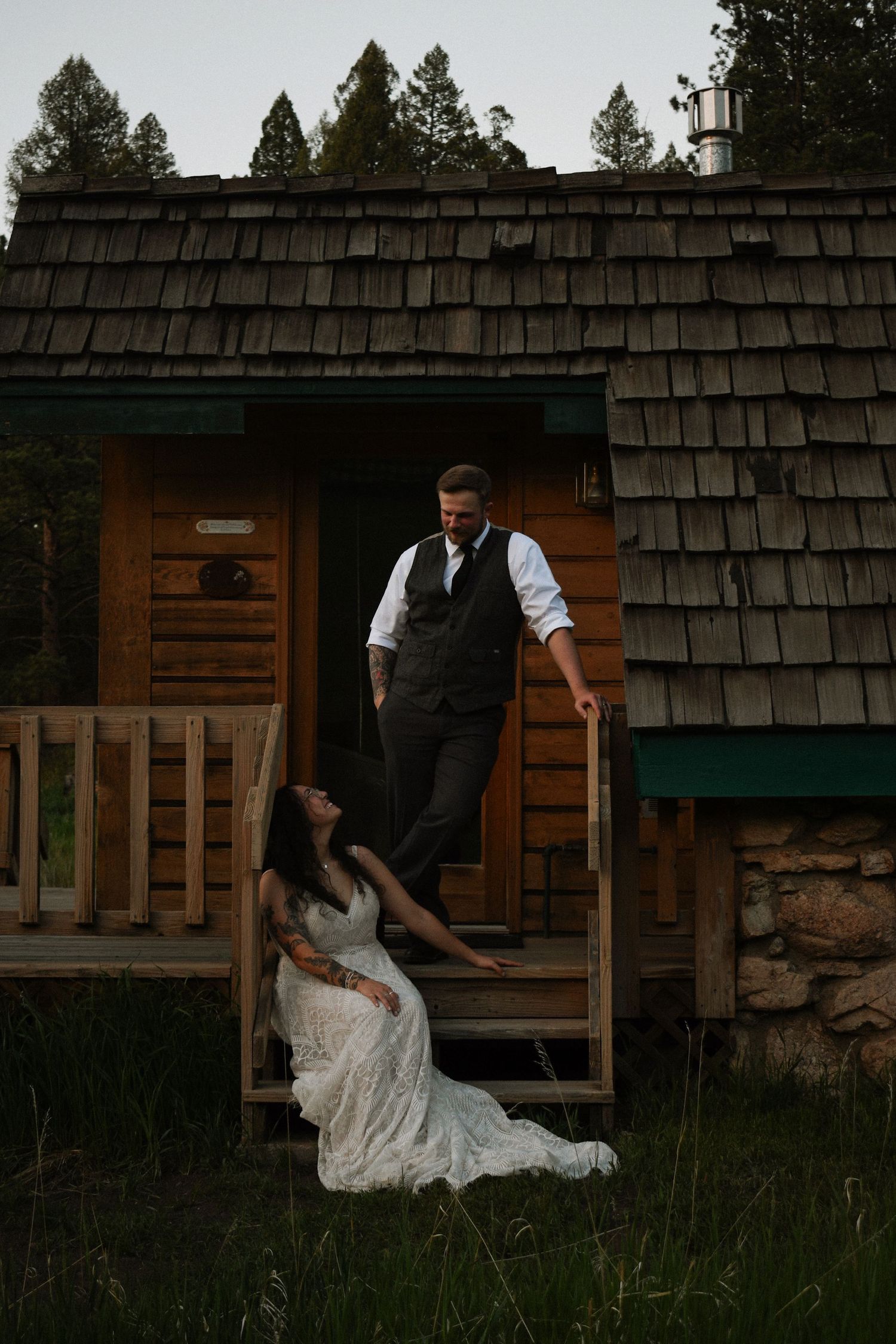 Bride in lace gown sitting on cabin steps gazing up at groom in vest at rustic mountain wedding.