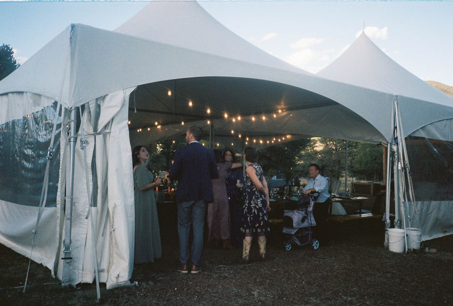 Guests mingle under a white tent with string lights at an outdoor evening event or wedding reception.