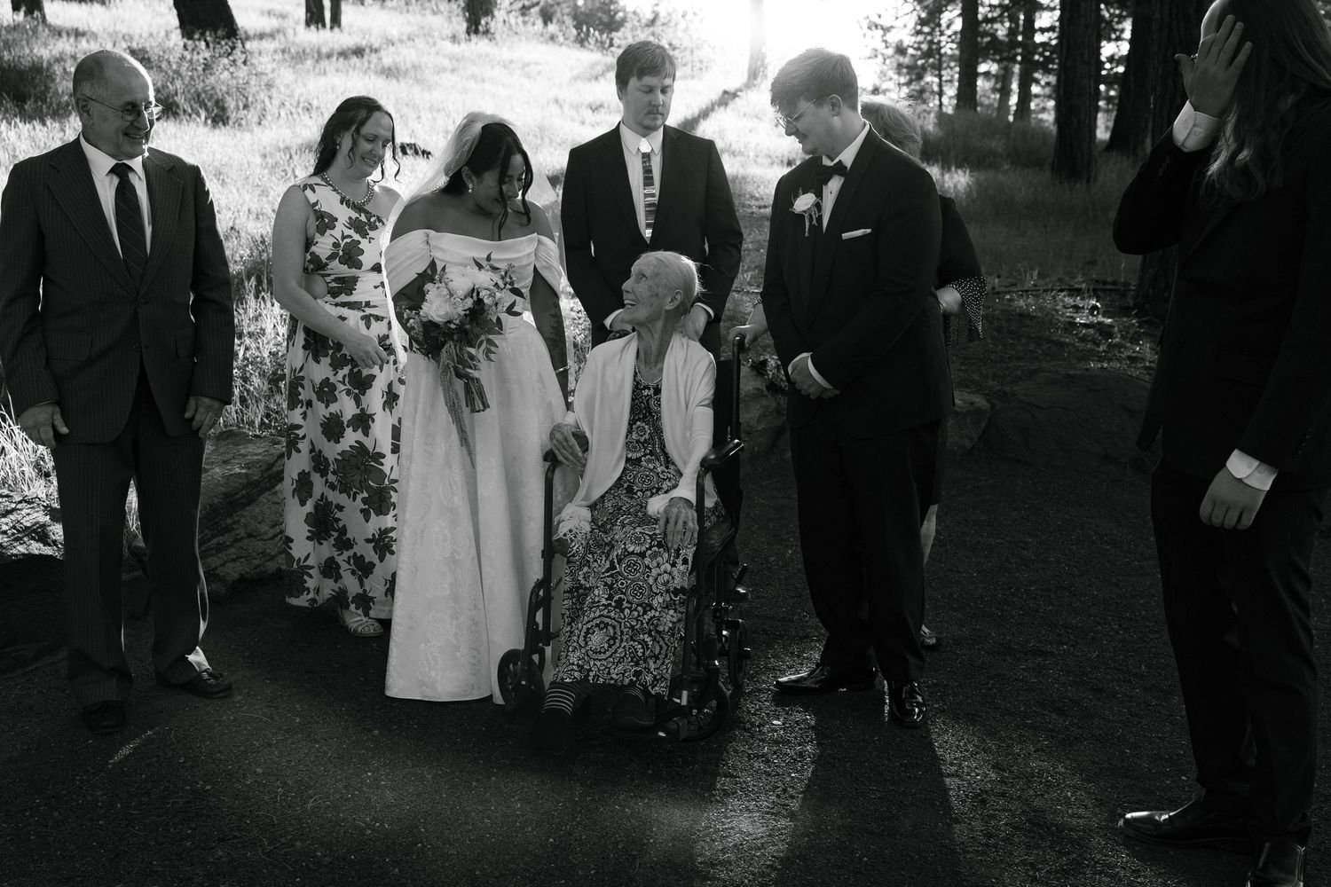 Black and white wedding photo of bride with bouquet, groom, and elderly woman in wheelchair surrounded by family outdoors.