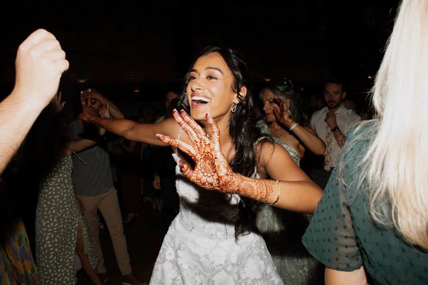 Bride with henna hands dancing joyfully at wedding reception with guests in background at night.