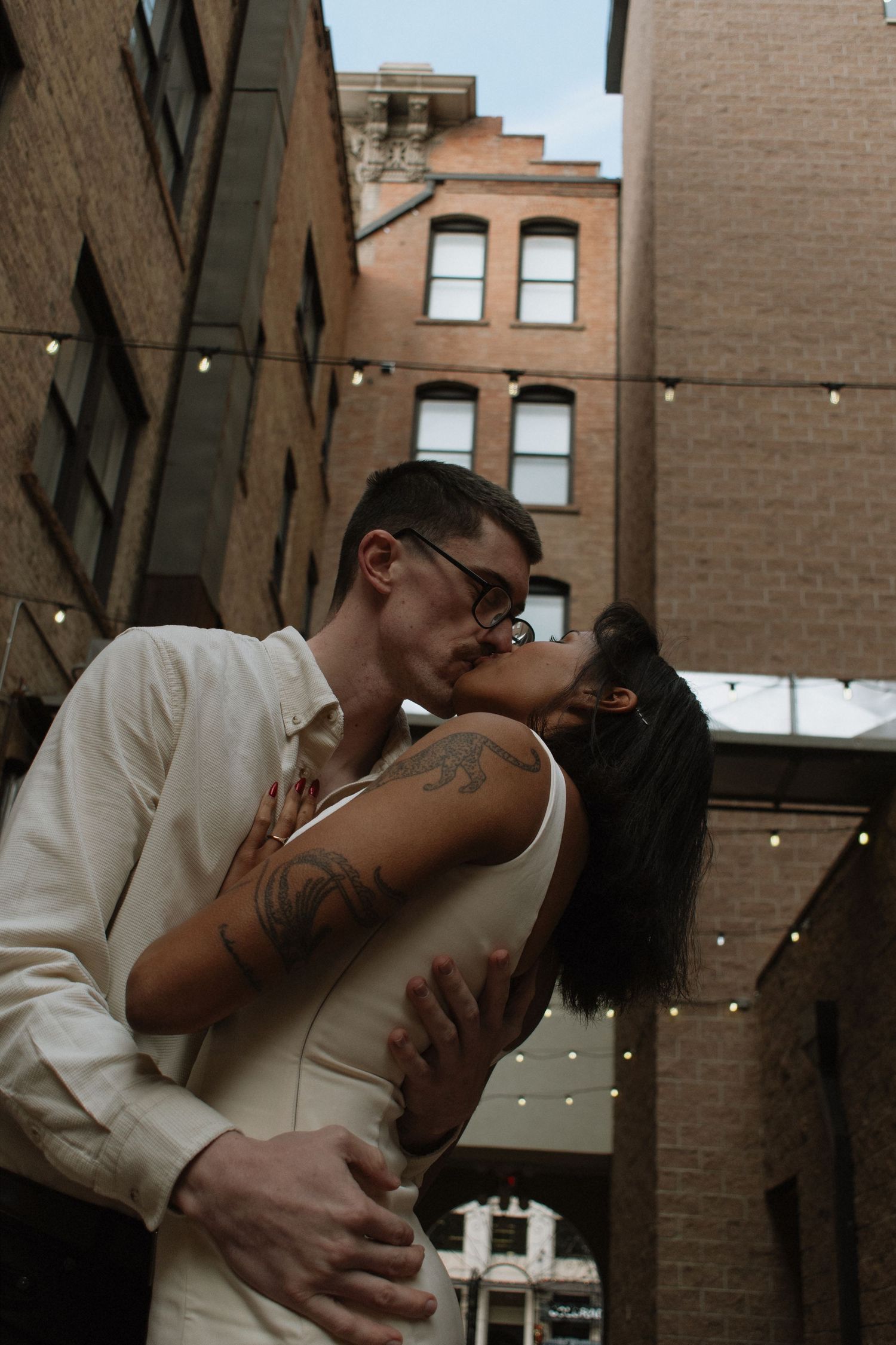 Couple sharing romantic kiss in urban alleyway with string lights and brick buildings.