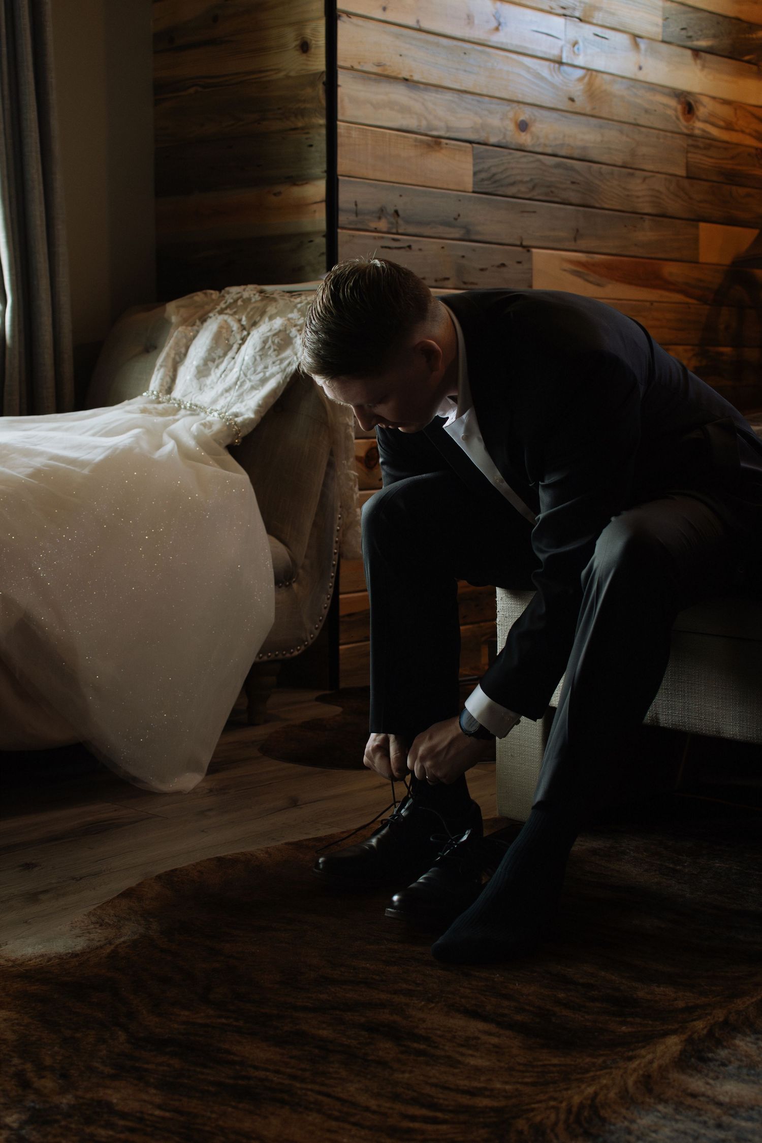 Groom in dark suit tying black dress shoes on wedding day, sitting beside bridal gown in rustic wood-paneled room.