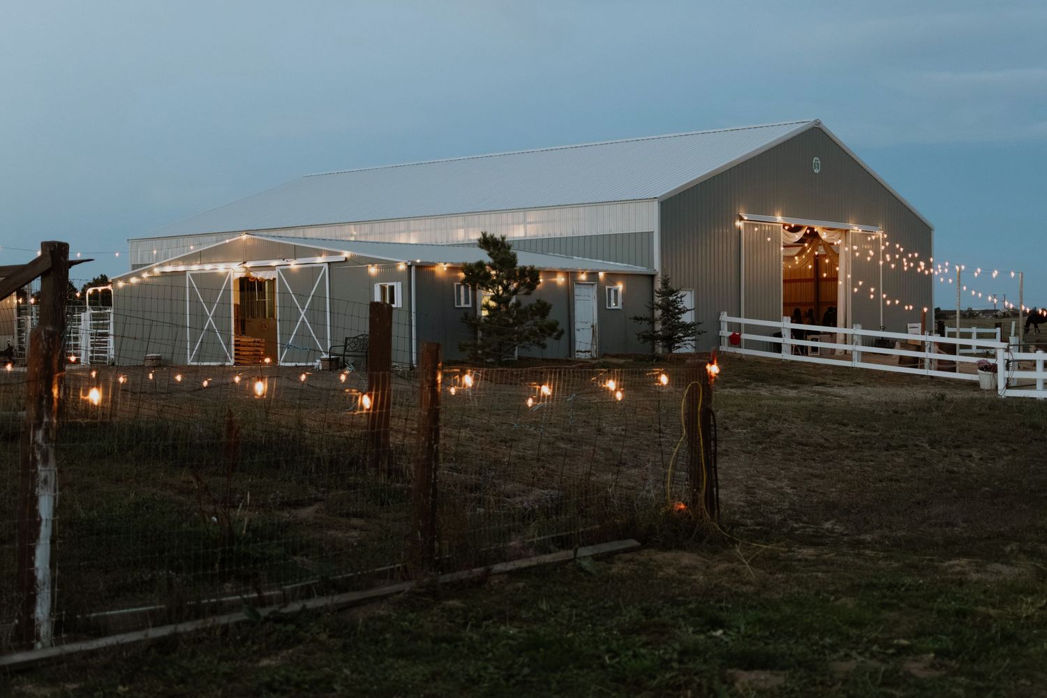 A rustic barn venue decorated with warm string lights at dusk, surrounded by a white fence on a rural farm property.