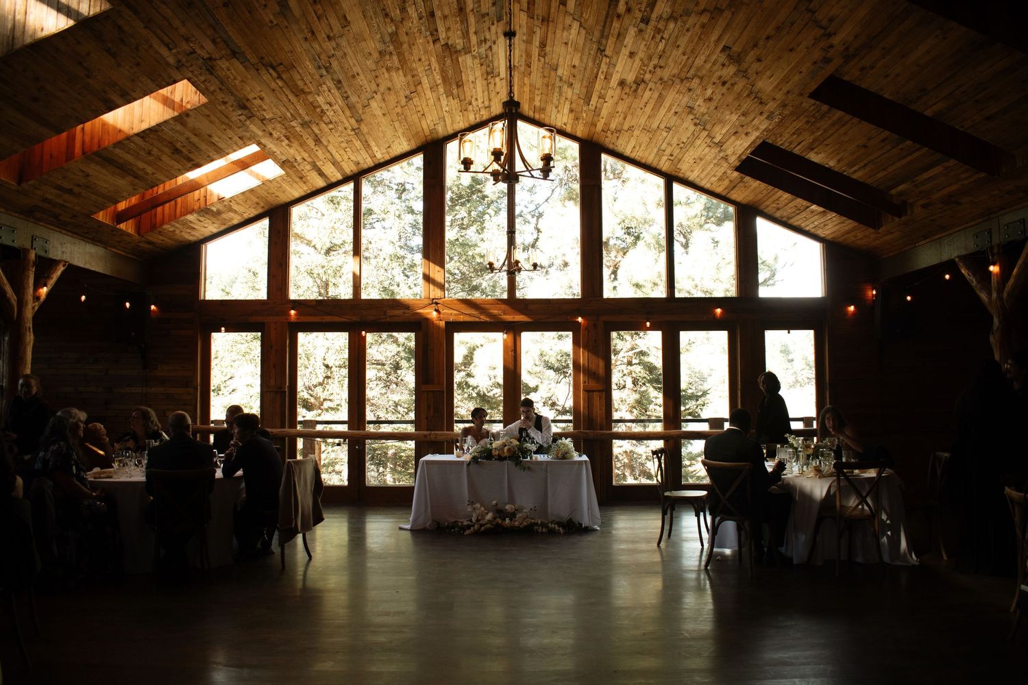Elegant barn wedding reception with dramatic floor-to-ceiling windows, chandelier, and head table silhouetted against natural light.