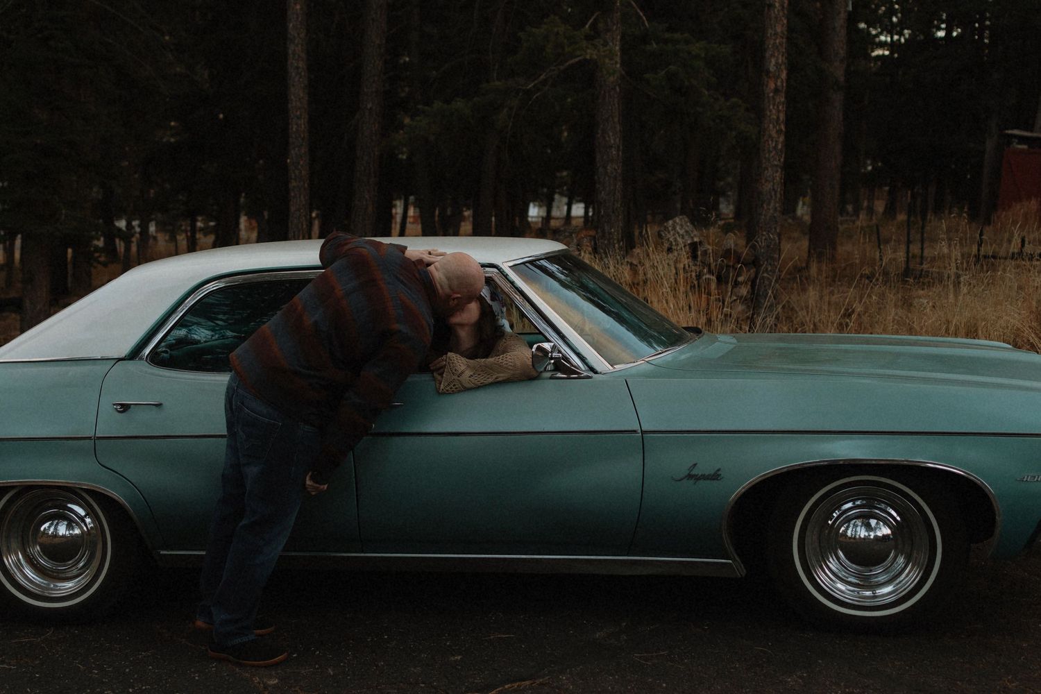 Man leaning into vintage teal classic car in a forest setting. Couple kissing