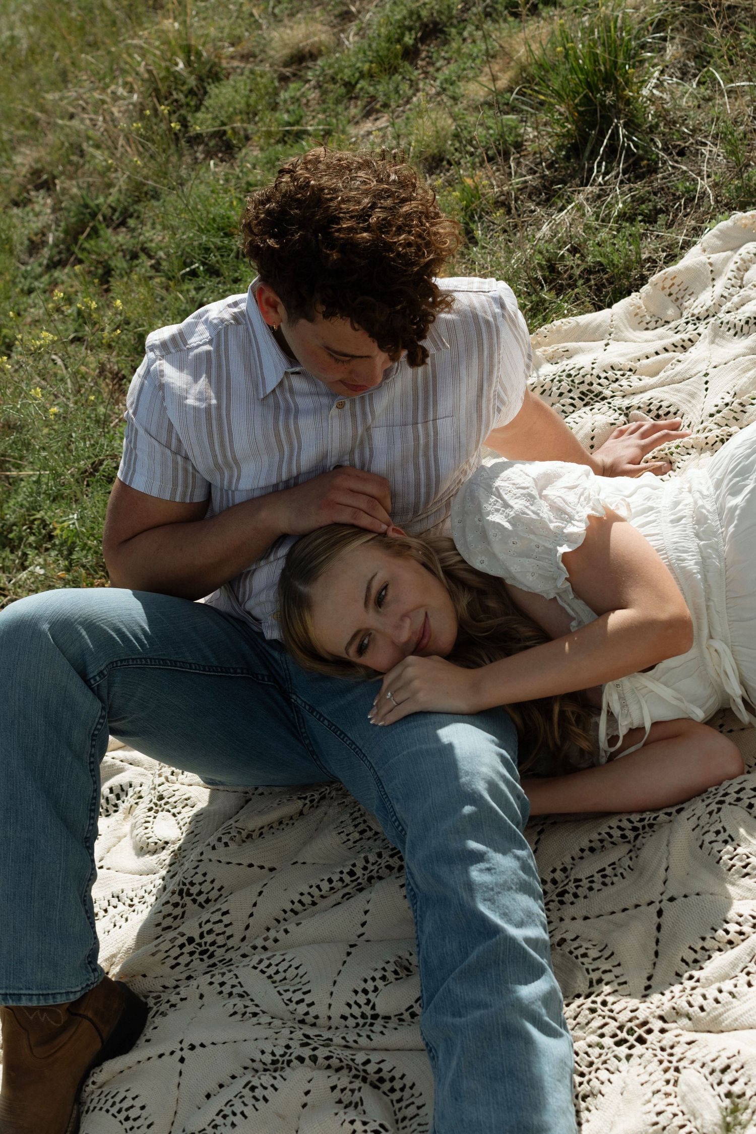 A couple relaxes on a lace blanket outdoors, woman lying on man's lap in white dress and jeans.
