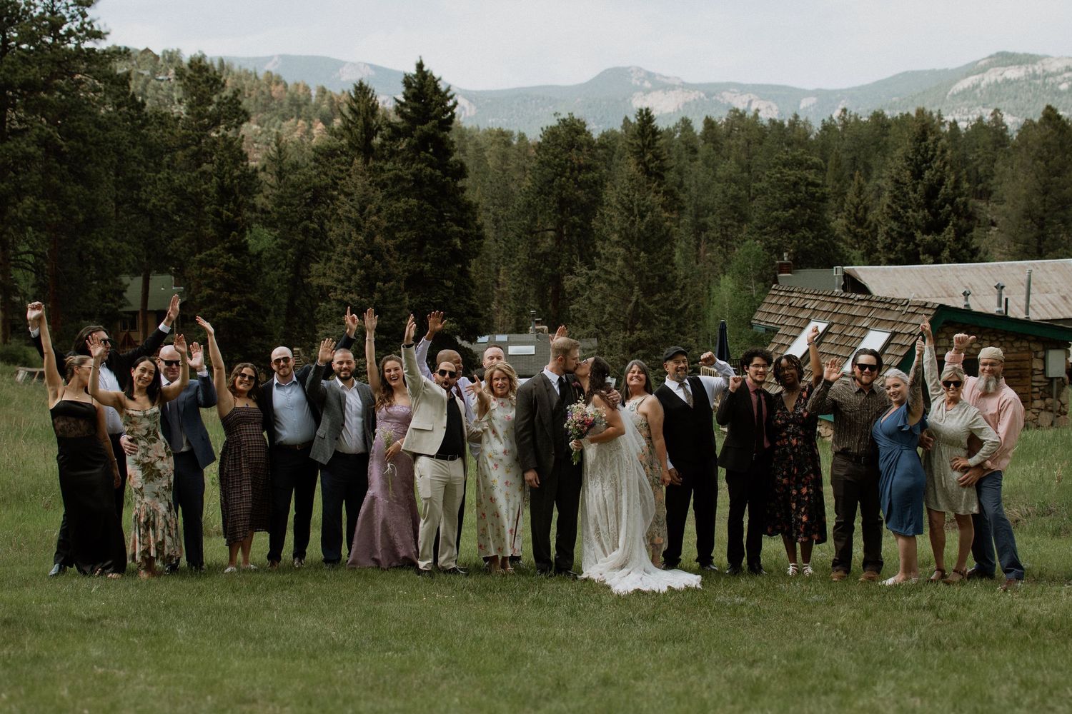 Large wedding group photo on green mountain meadow with forested peaks and rustic venue in background.