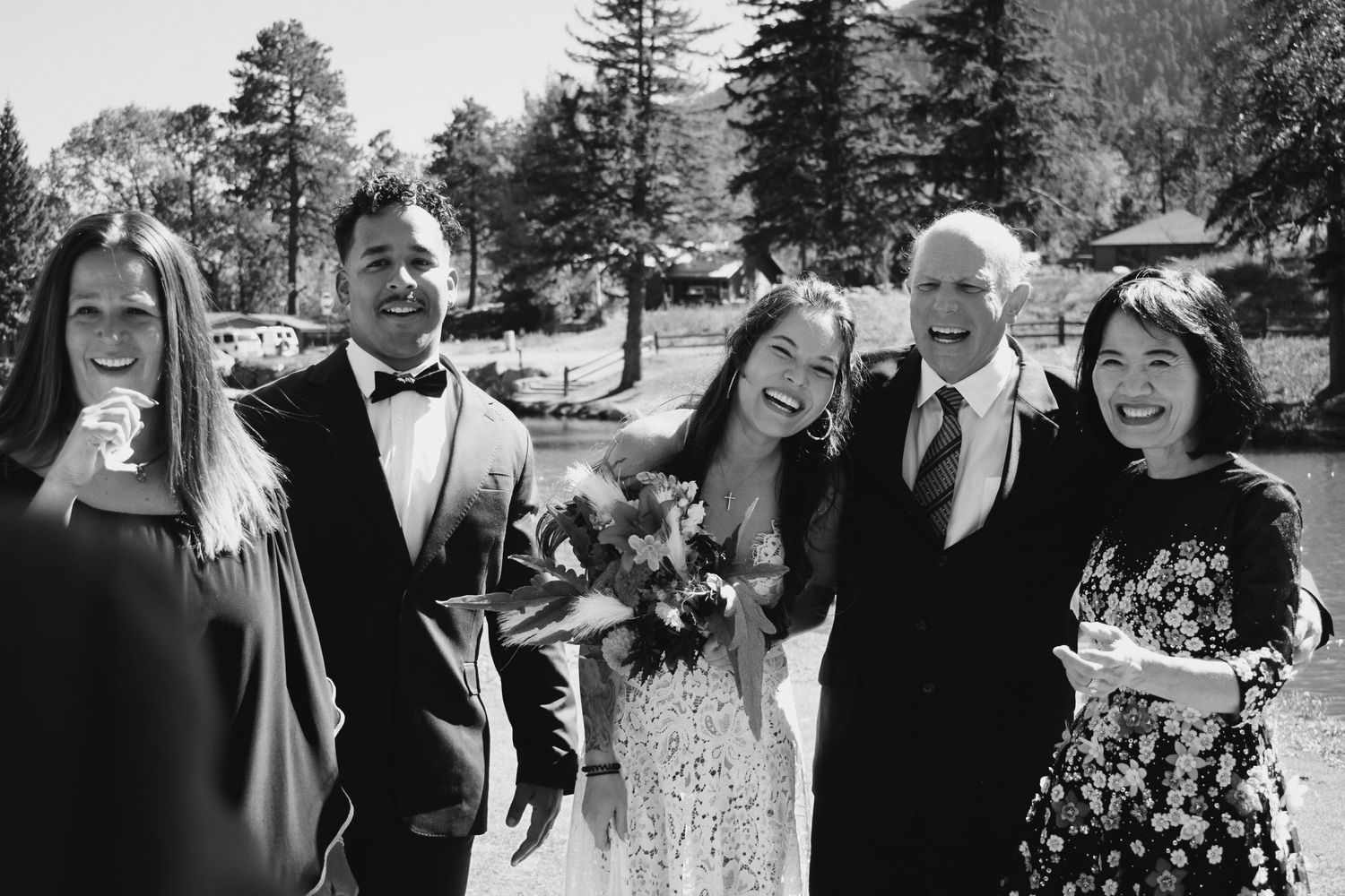 Black and white wedding photo of smiling bride and groom with family outdoors among tall trees.