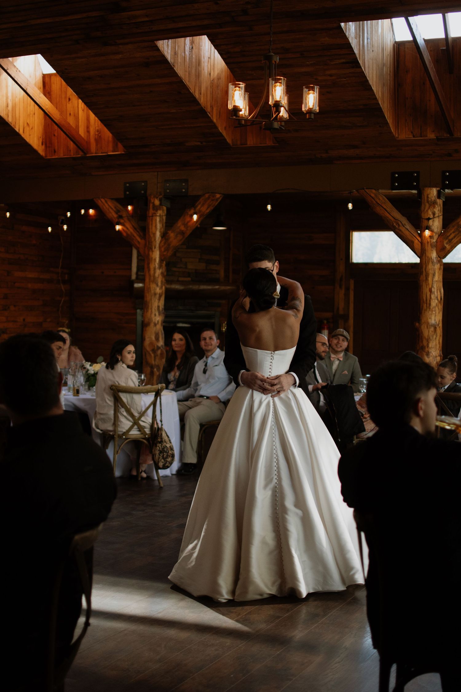 Bride and groom share their first dance in a rustic wooden barn venue with warm string lights and guests seated around them.