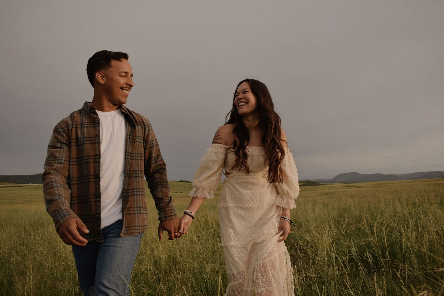 Couple holding hands and laughing in a green field under a dramatic cloudy sky, wearing casual and bohemian outfits.