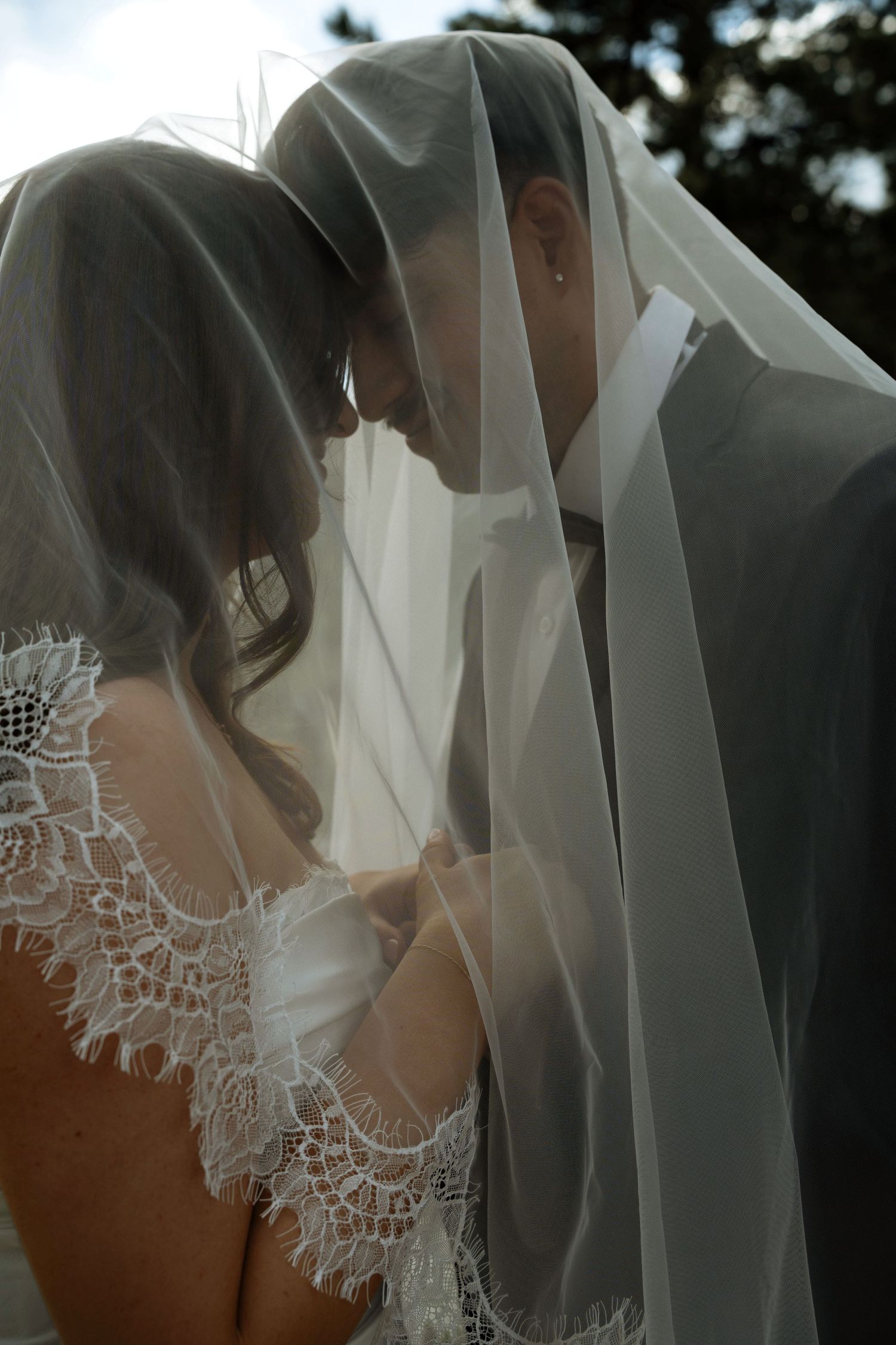 Bride and groom share an intimate moment under a sheer wedding veil, bride wearing lace dress.