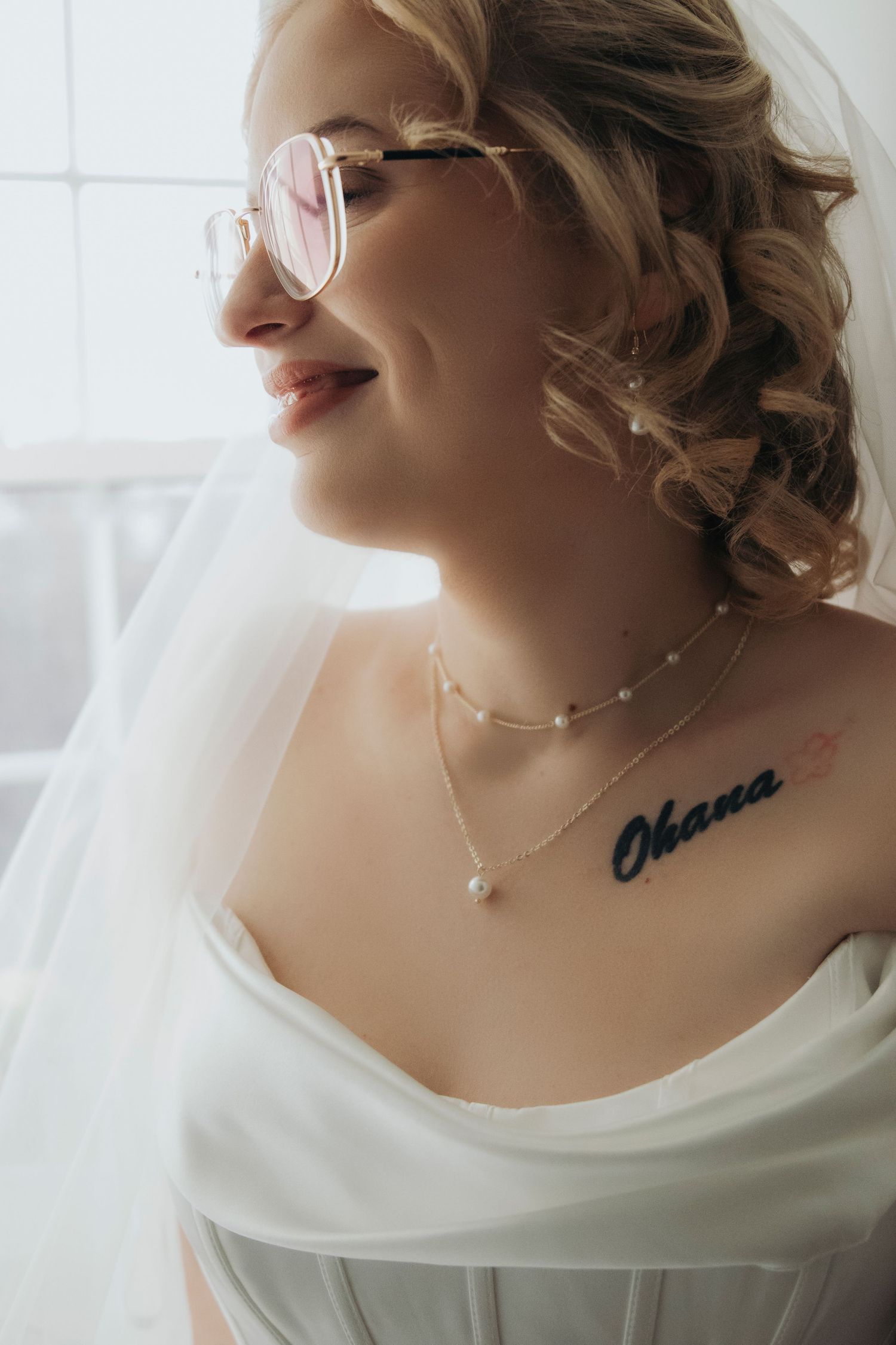 Smiling bride in pink sunglasses with Ohana chest tattoo, pearl necklace, and white veil on wedding day.