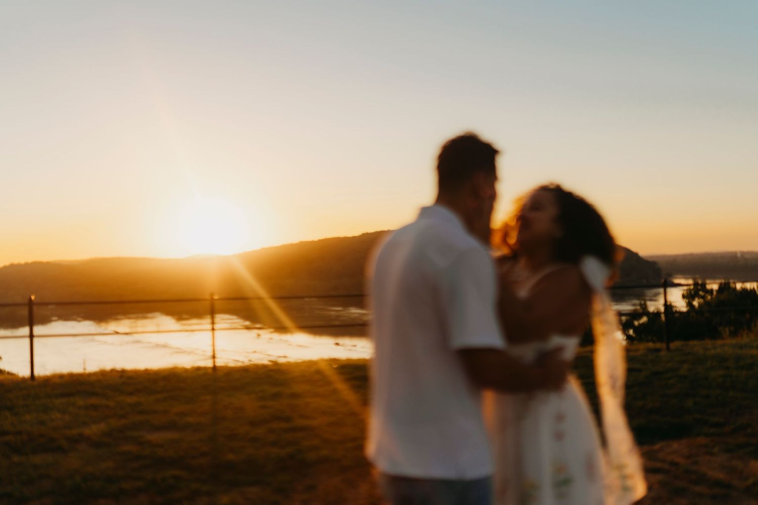 A couple embraces at sunset on a hilltop overlooking a scenic landscape with golden light in the background.