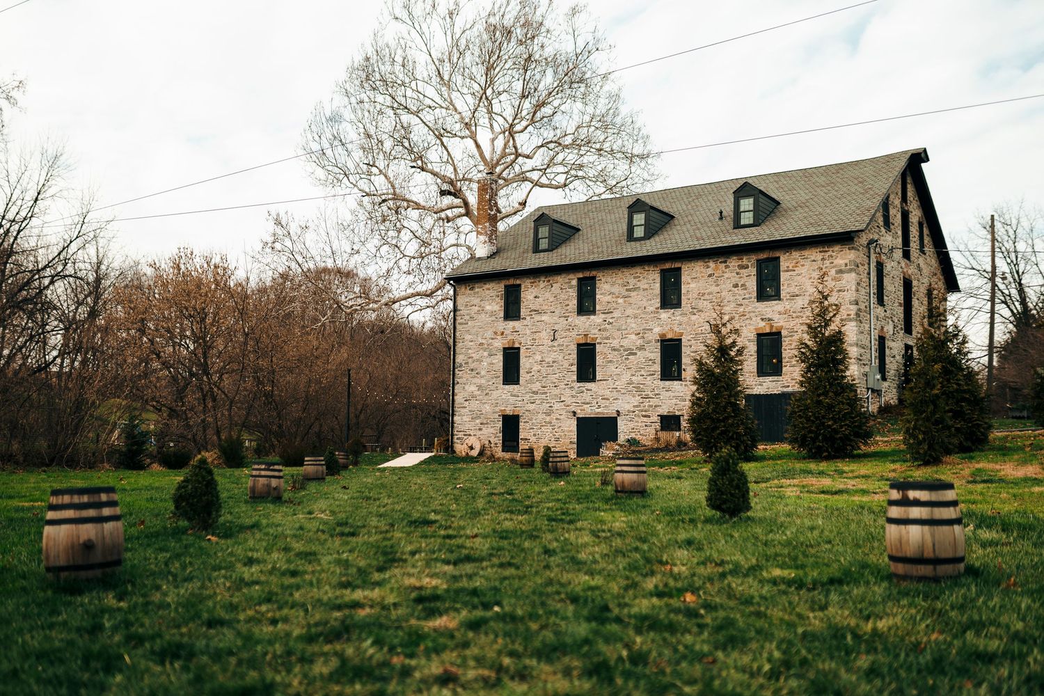 Historic stone mansion with dormer windows sits on a green lawn with wooden barrels and shrubs scattered across the property.