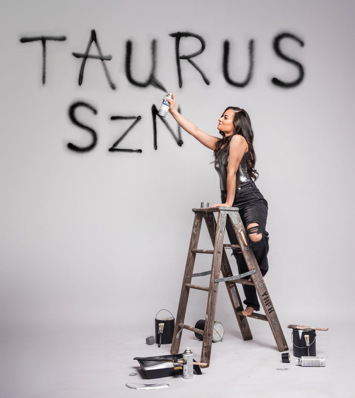 A creative photo of someone on ladder writing 'Taurus SZN' on white wall with paint supplies.