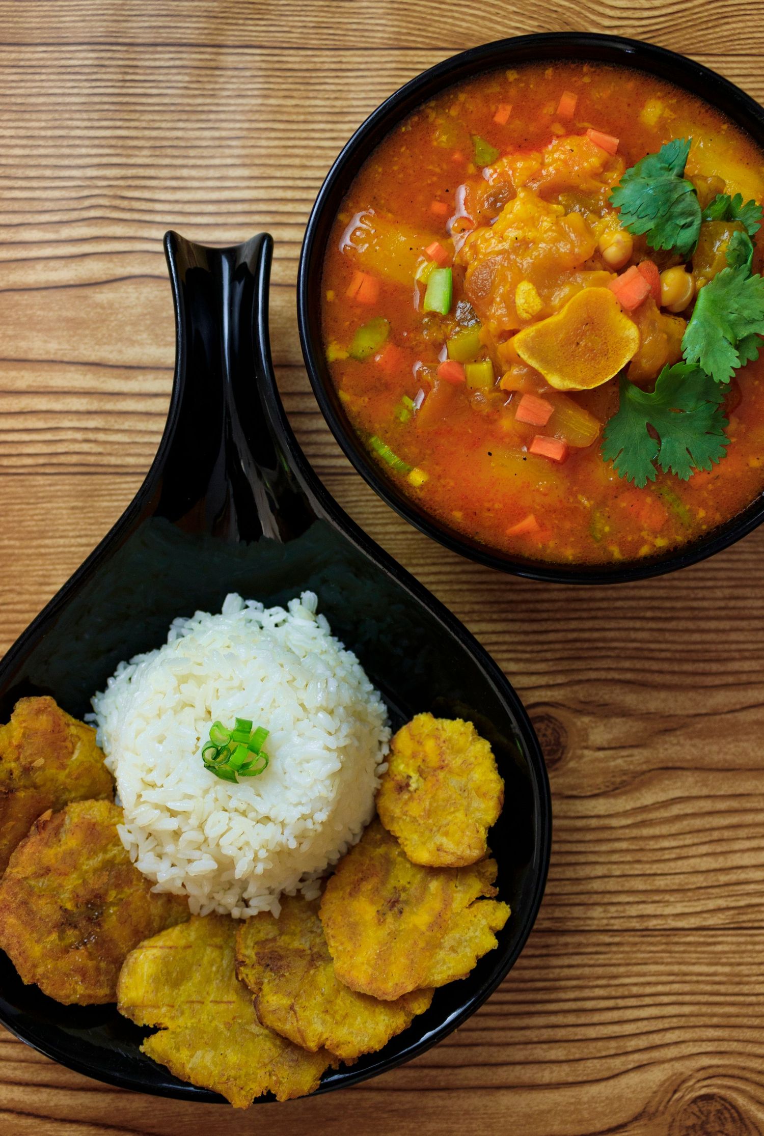 Caribbean curry stew with plantains and rice served in black dishes on a wooden table.