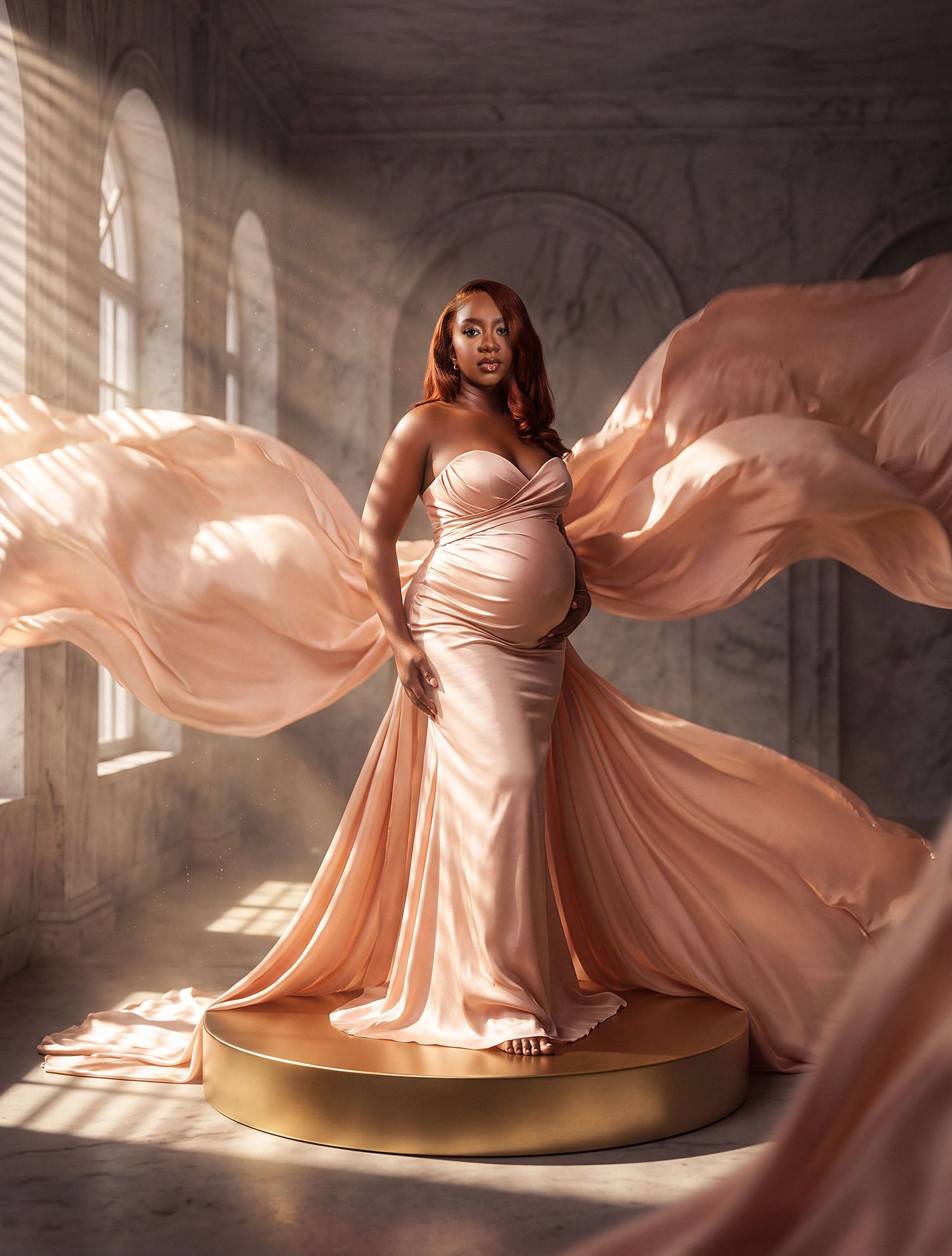 Pregnant woman in elegant peach satin gown with flowing fabric wings standing on gold platform in sunlit marble room.
