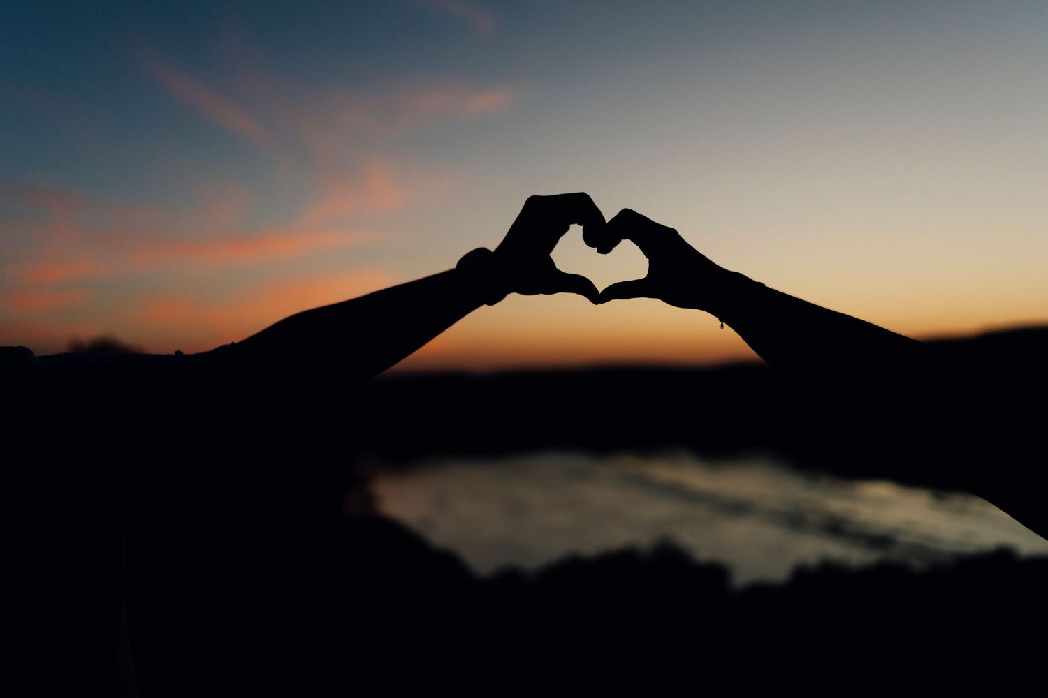 Silhouetted hands forming a heart shape against a dramatic sunset sky over mountain landscape.