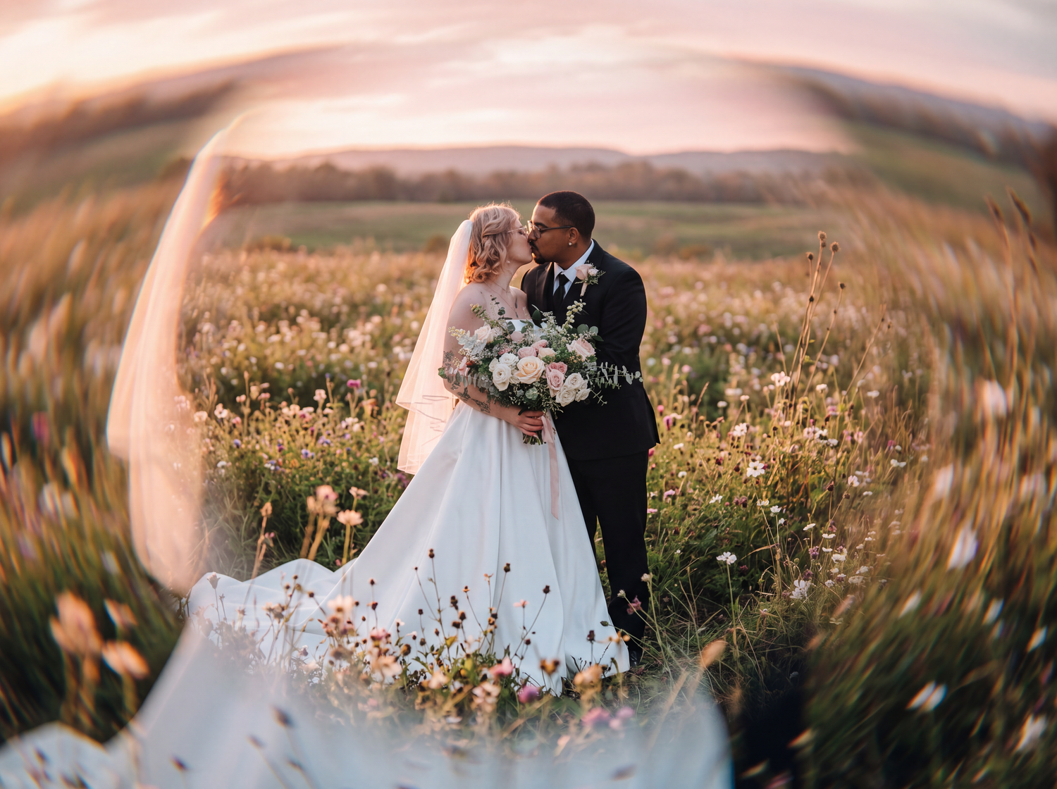 Bride and groom kissing in a wildflower field at sunset, with flowing veil and bouquet in romantic wedding photo.