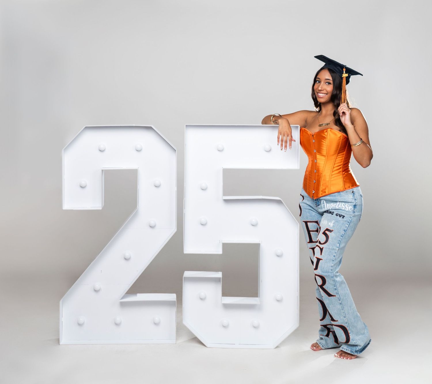 Graduate in orange top and decorated jeans poses next to large illuminated number 25 against grey background.