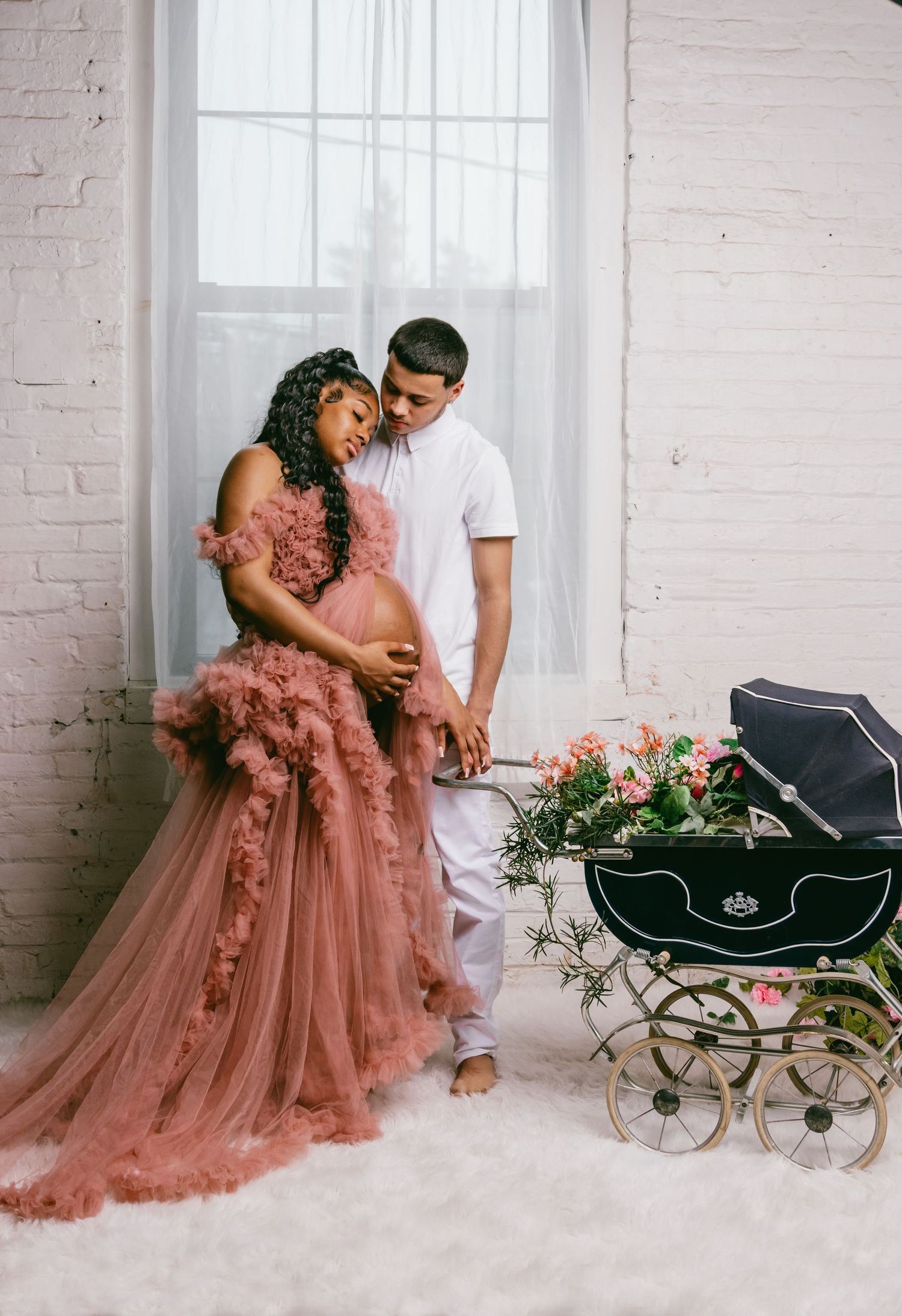 A romantic maternity photo with couple in dusty rose gown near vintage pram with floral arrangements.