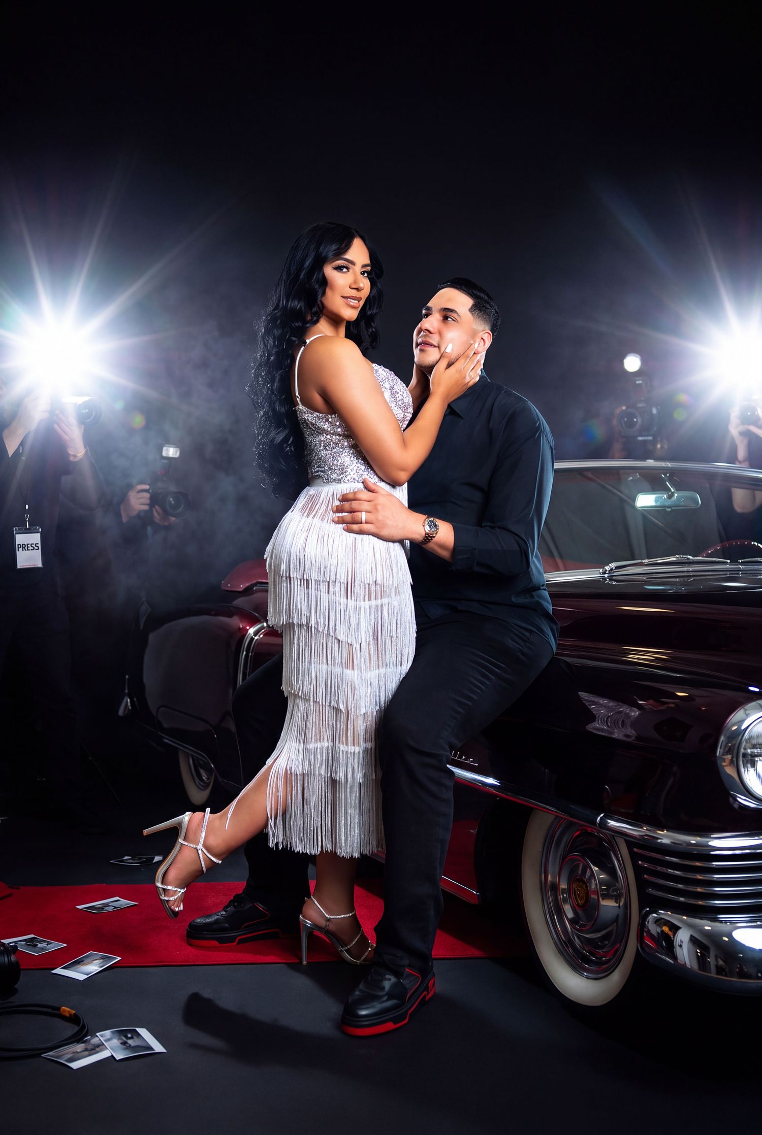 A glamorous couple poses on a red carpet beside a vintage car, with paparazzi flashing cameras in the background.