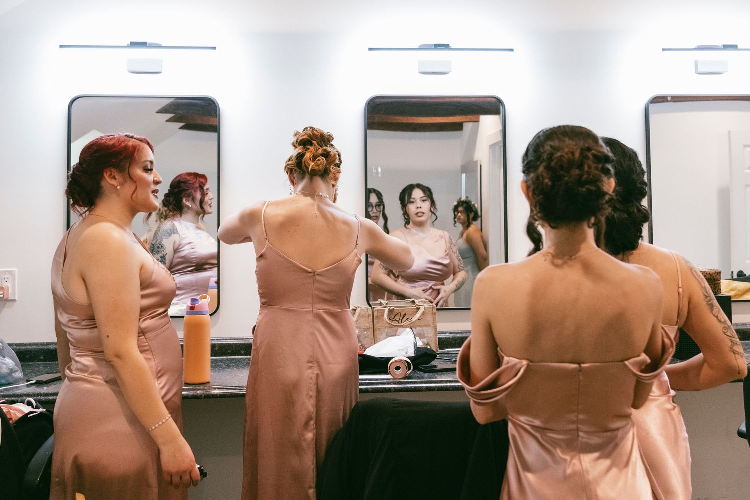 Bridesmaids in blush pink gowns helping each other get ready in a mirrored dressing room before a wedding.
