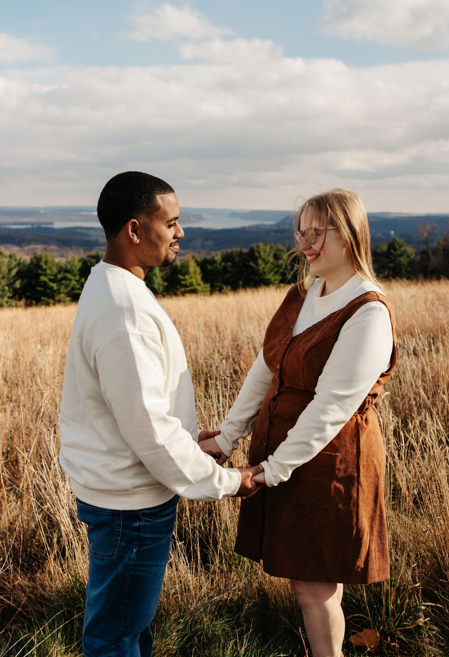 A couple shares a playful moment in a golden field during sunset, wearing coordinating white and brown outfits.