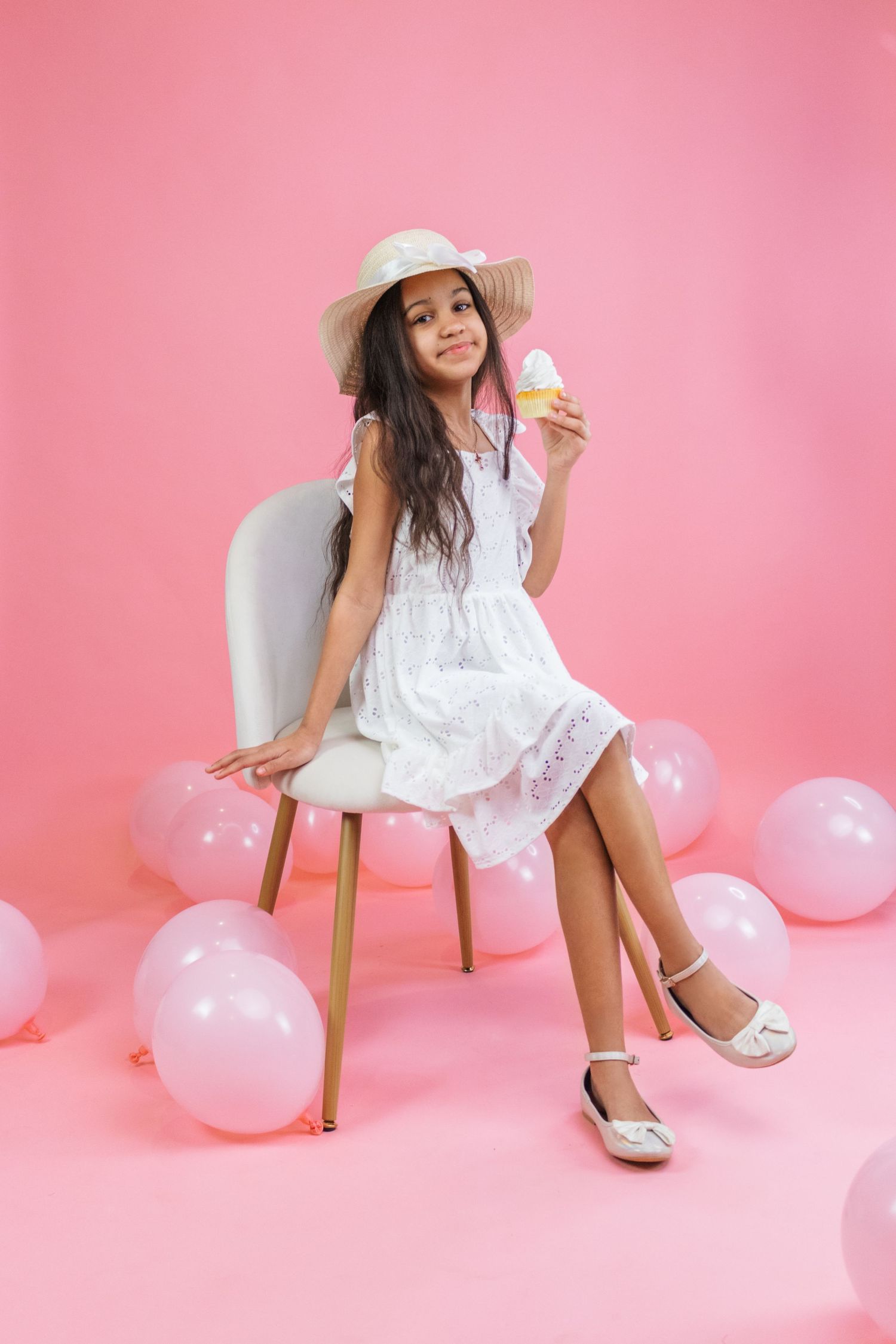 A summery portrait with white dress, sun hat and pink balloons in a studio setting.