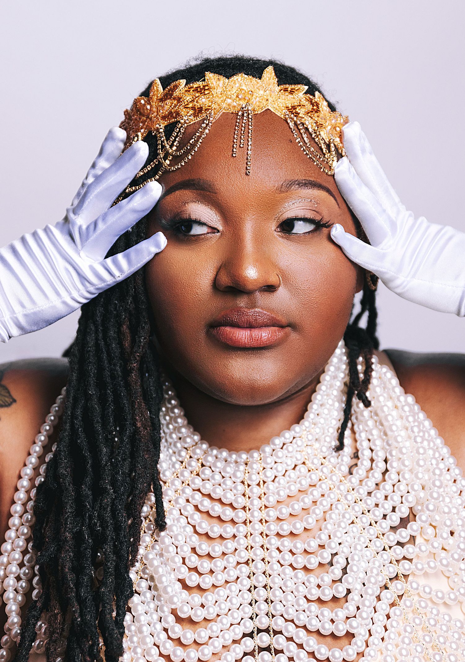 Person wearing an ornate gold crown and pearl necklace with gloved hands touching their face.