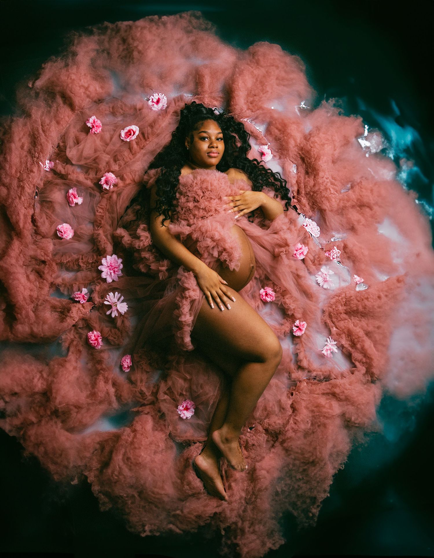 Ethereal maternity photo with pink smoke effect and flower petals creating dreamy atmosphere.