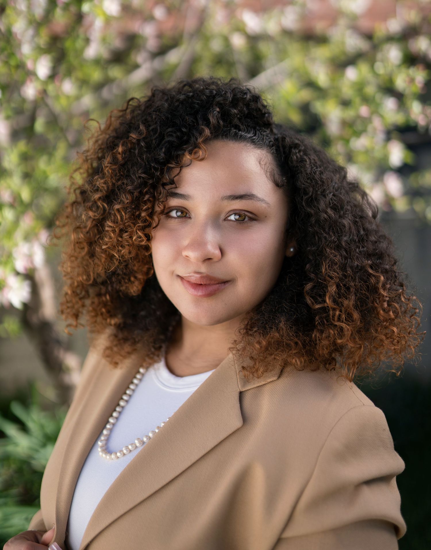 Young professional woman with curly hair wearing a tan blazer and pearl necklace poses outdoors.