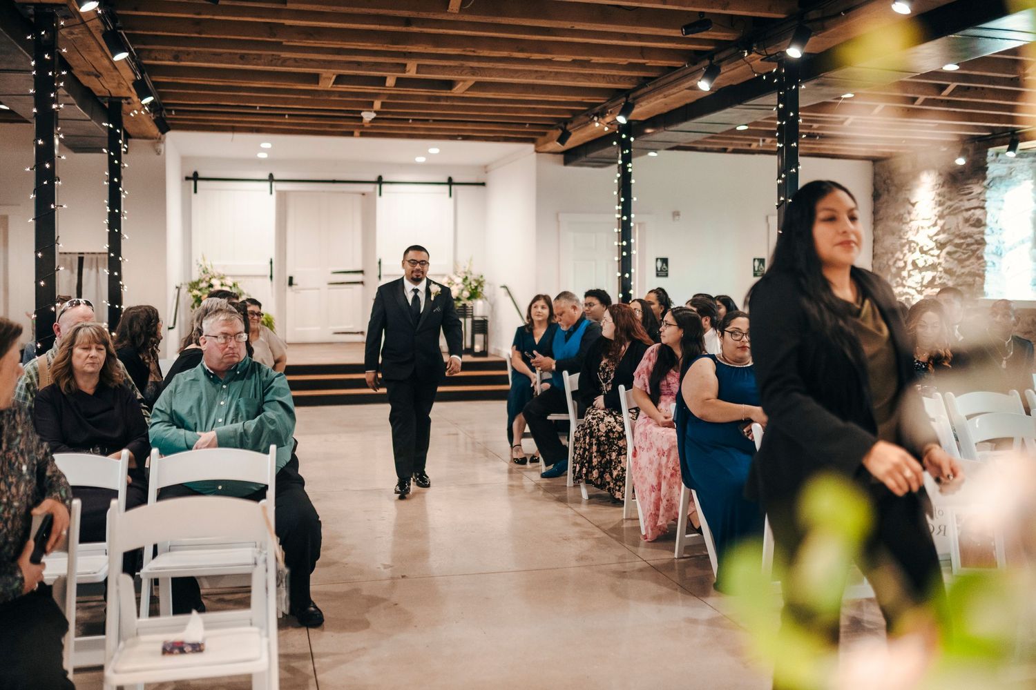 A groom walks down the aisle at an indoor wedding ceremony with guests seated in white chairs on both sides.