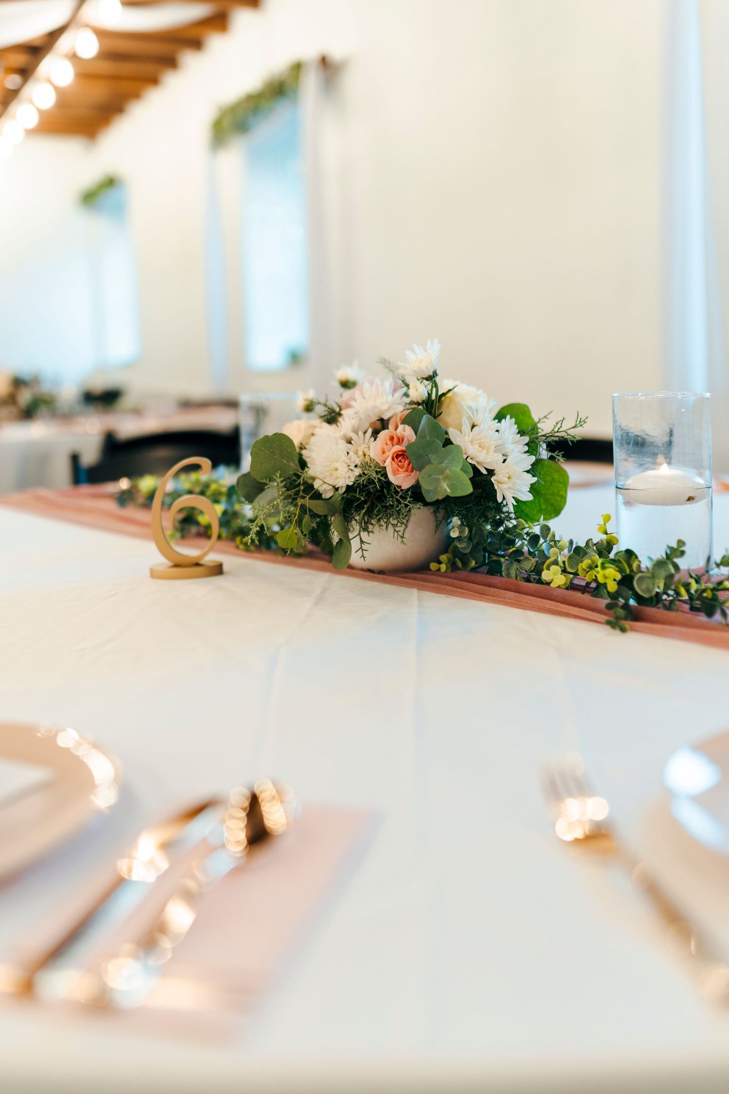 Elegant wedding reception table with white floral centerpiece, greenery runner, blush napkins, and gold accents.