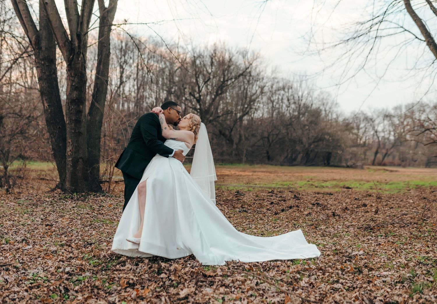Bride and groom sharing romantic dip kiss among autumn fallen leaves beneath bare trees.