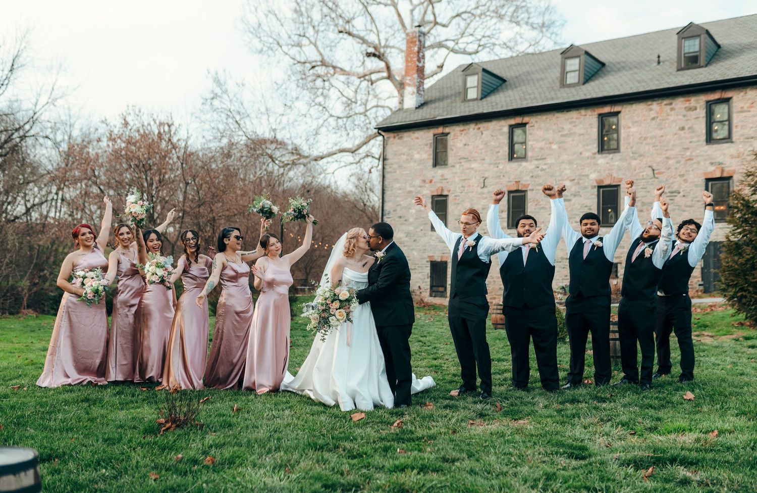 Wedding party celebrating outdoors, bridesmaids in pink dresses, groomsmen in suits, bride and groom kissing.
