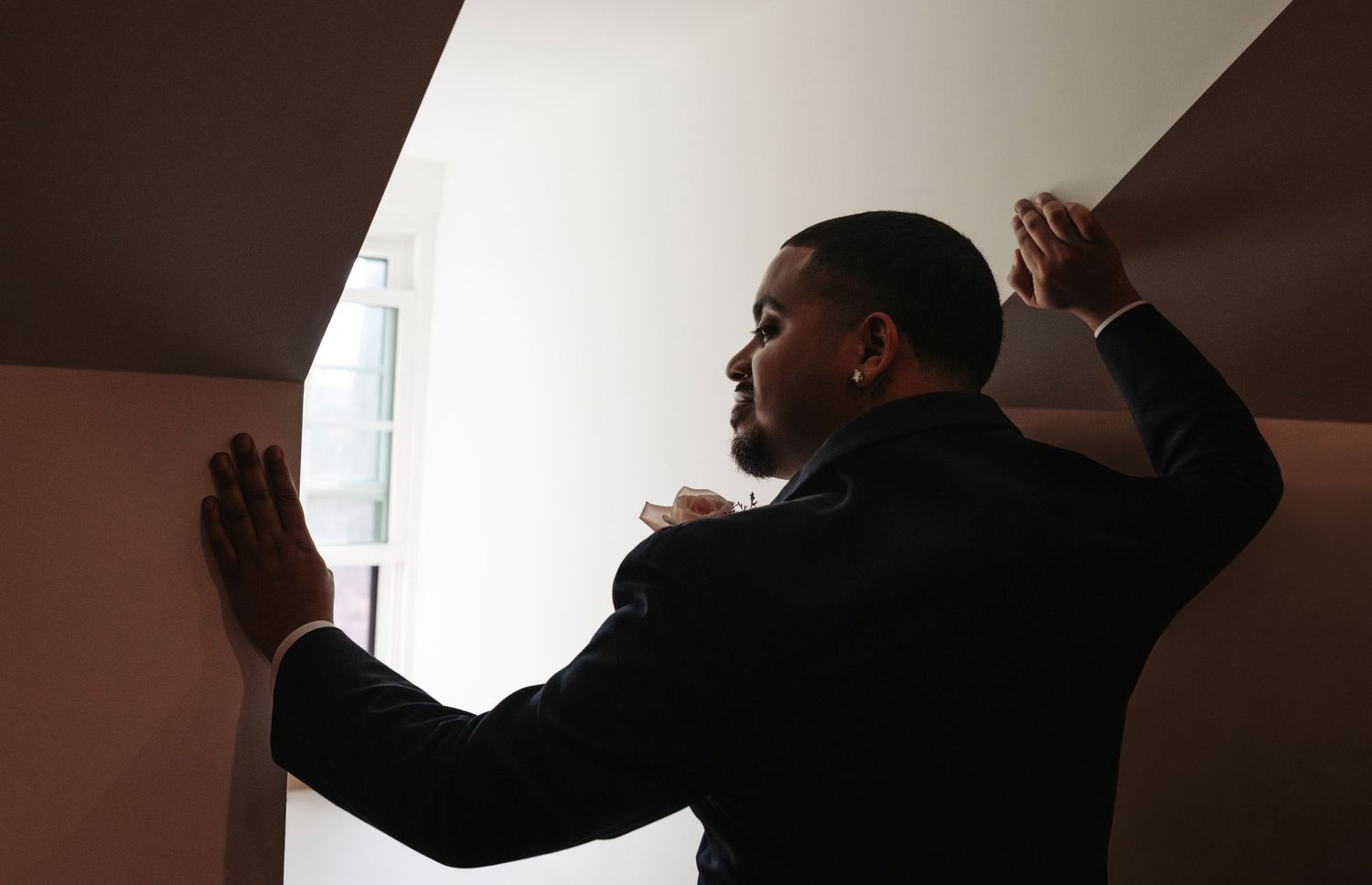 A man in a suit stands with hands pressed against walls in a narrow corridor, looking out a window.