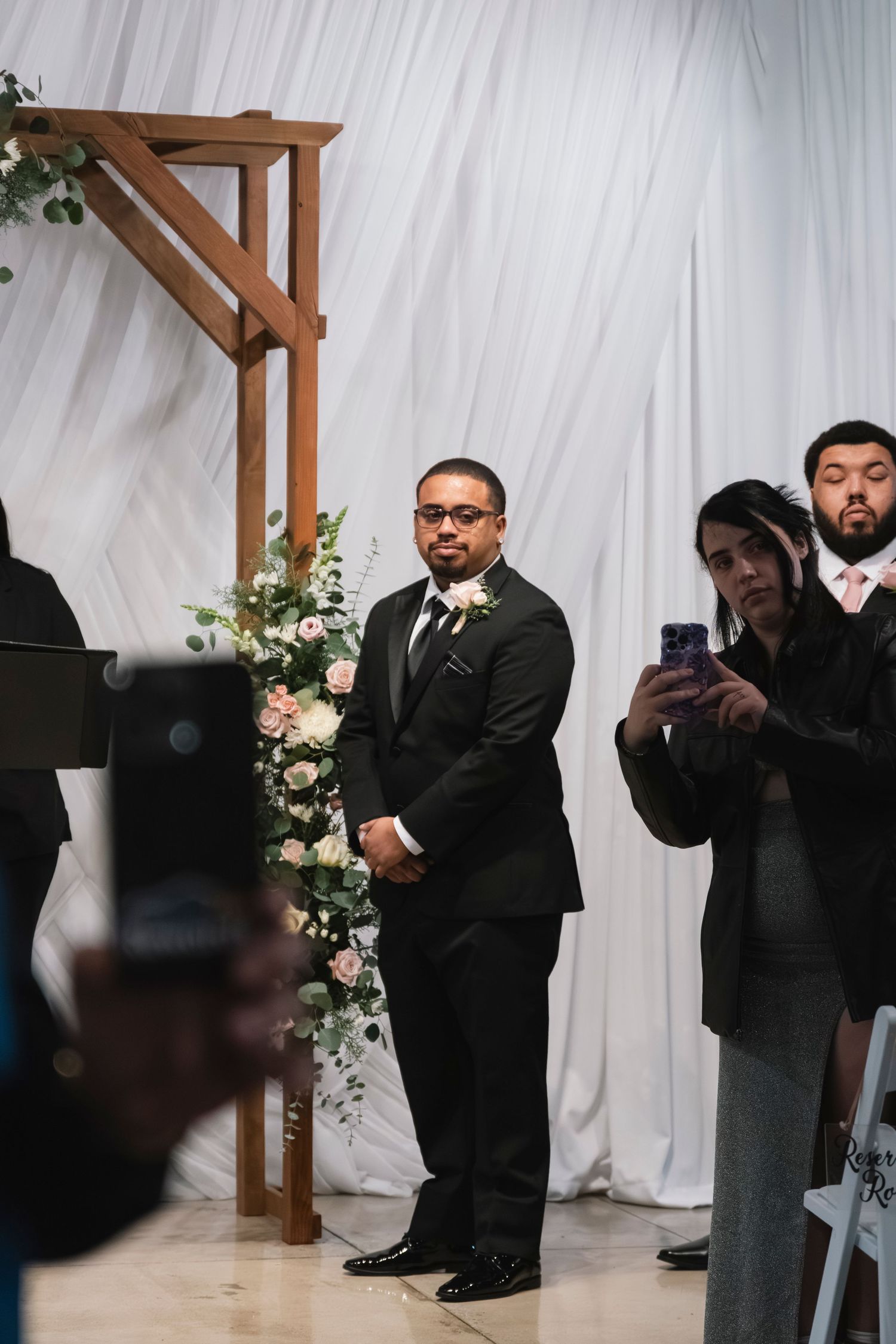 Groom in black tuxedo standing at floral wedding arch awaiting bride during ceremony.