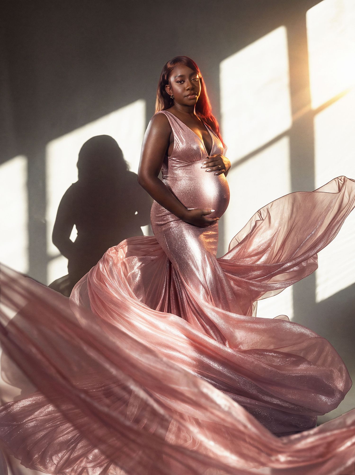 Pregnant woman in flowing rose gold gown poses gracefully in sunlit studio with dramatic shadow backdrop.