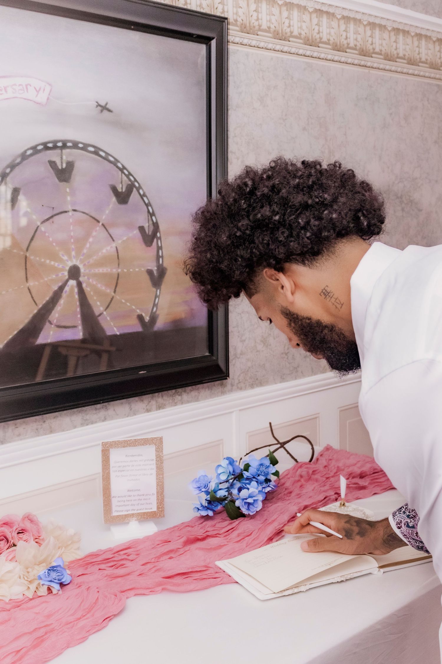 Person signing guestbook beside framed ferris wheel artwork and pink table runner.