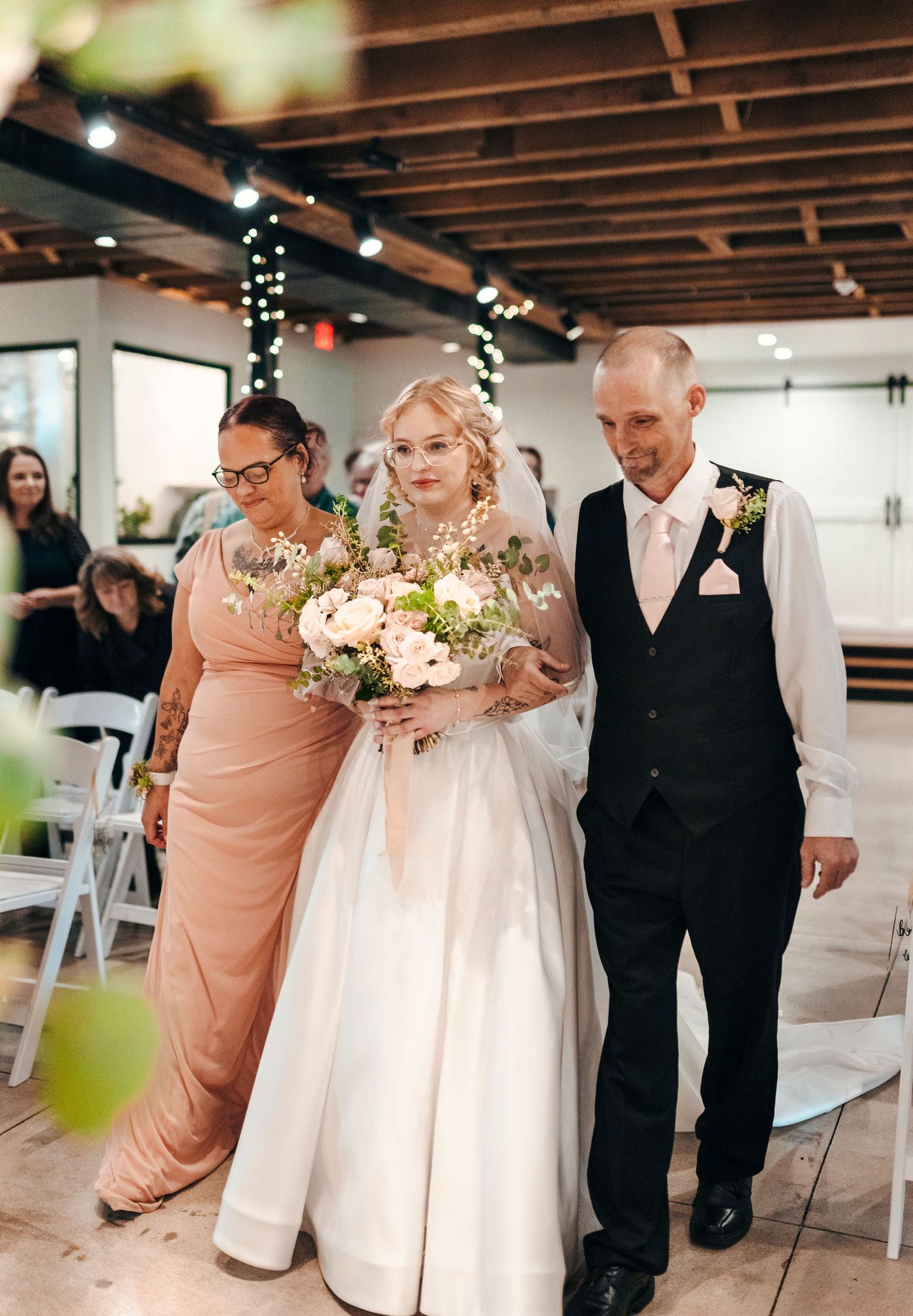Bride walks down the aisle with parents holding a pink floral bouquet at an indoor wedding ceremony.