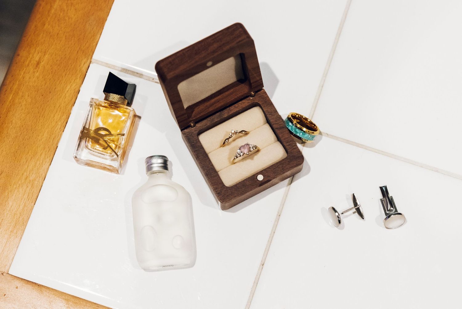 Wedding flat lay with gold rings in wooden box, perfume bottles, turquoise ring, and silver cufflinks on white surface.