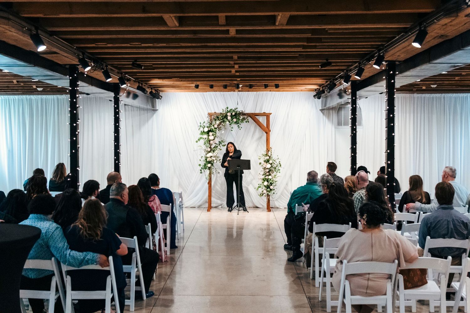 Indoor wedding ceremony with guests seated in white chairs facing an officiant at a floral arch altar.