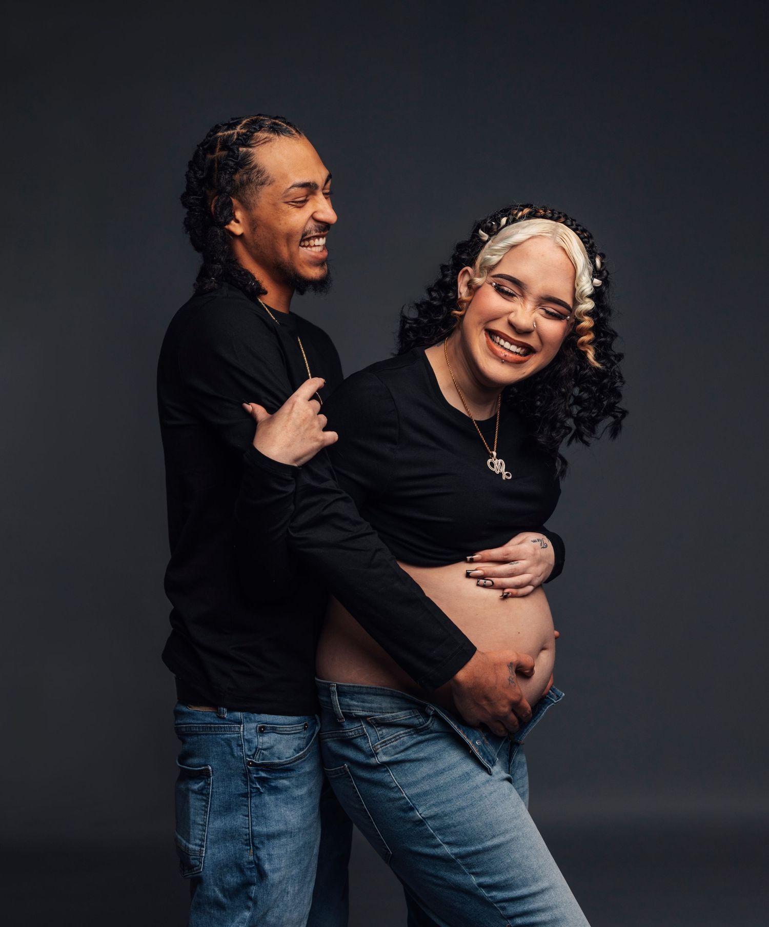 Expecting couple in casual wear sharing joyful moment during pregnancy photoshoot against dark background.