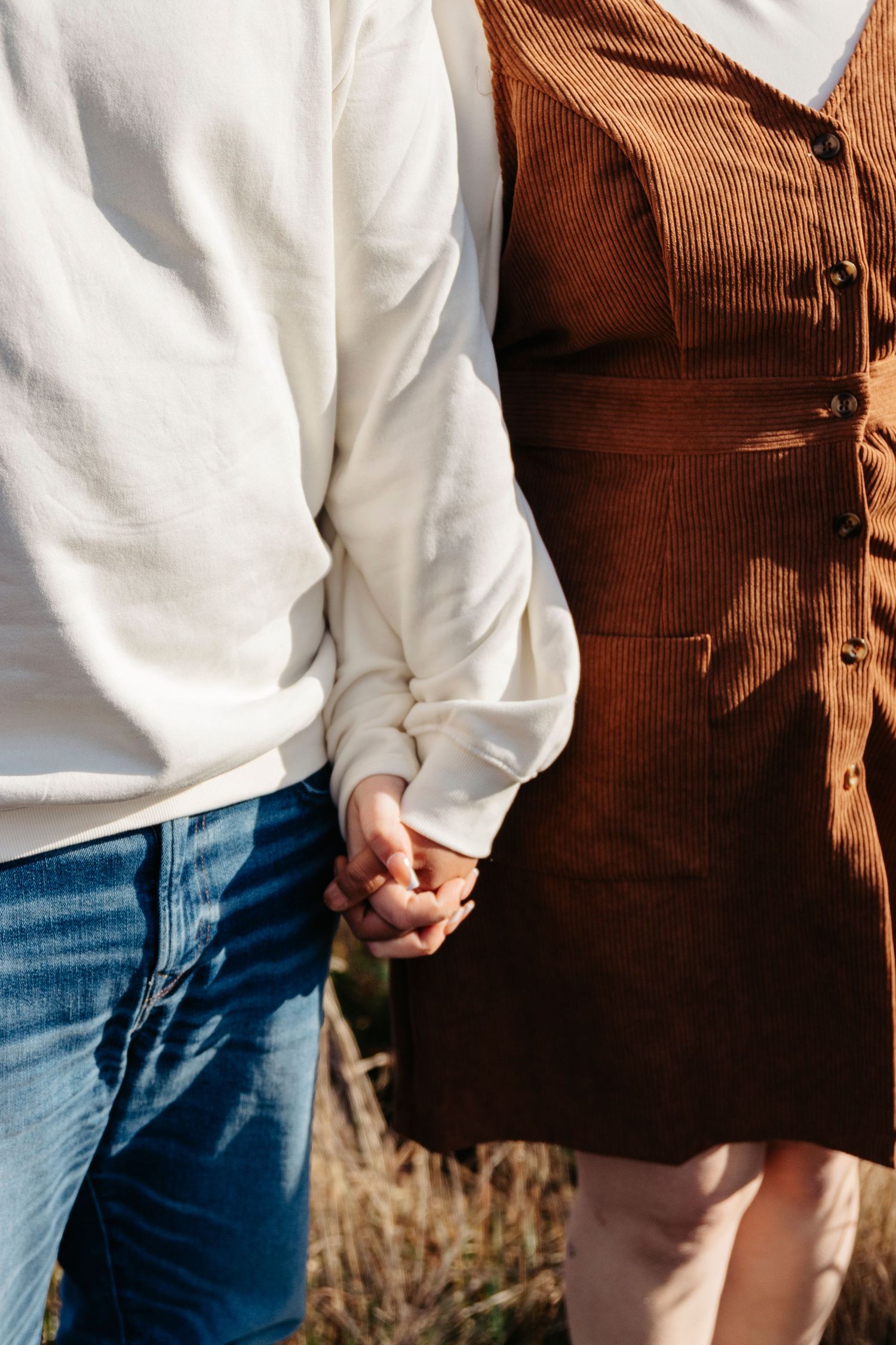Couple wearing brown corduroy vest and white shirt hold hands while walking through a field in autumn.