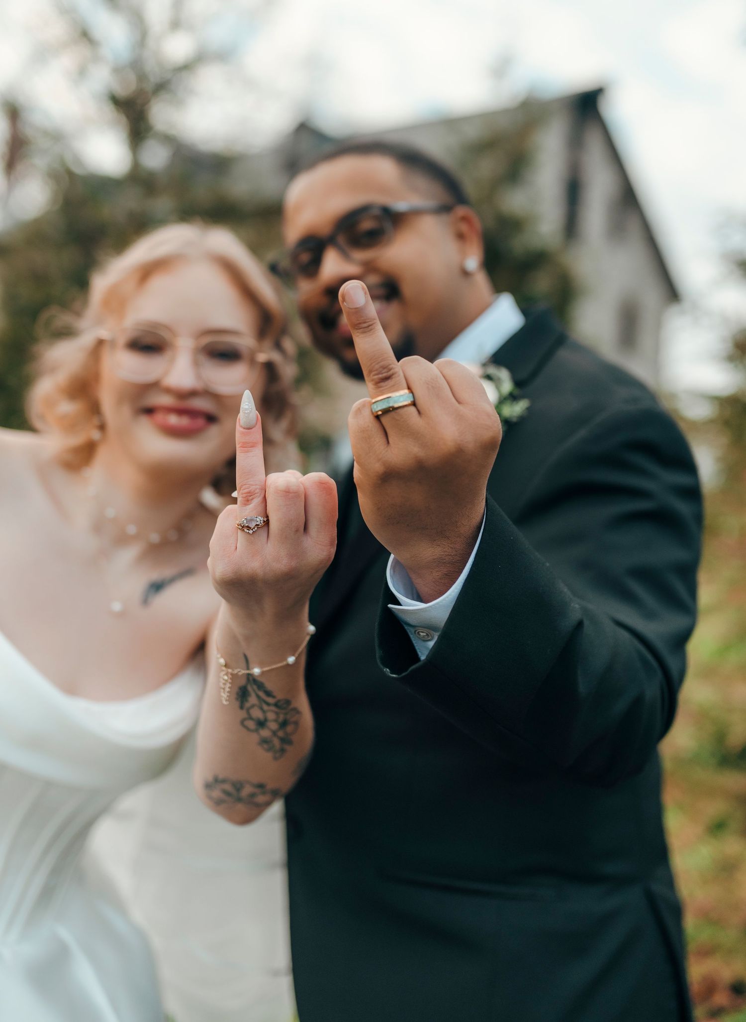 Newlywed couple smiling and showing off wedding rings on middle fingers outdoors.