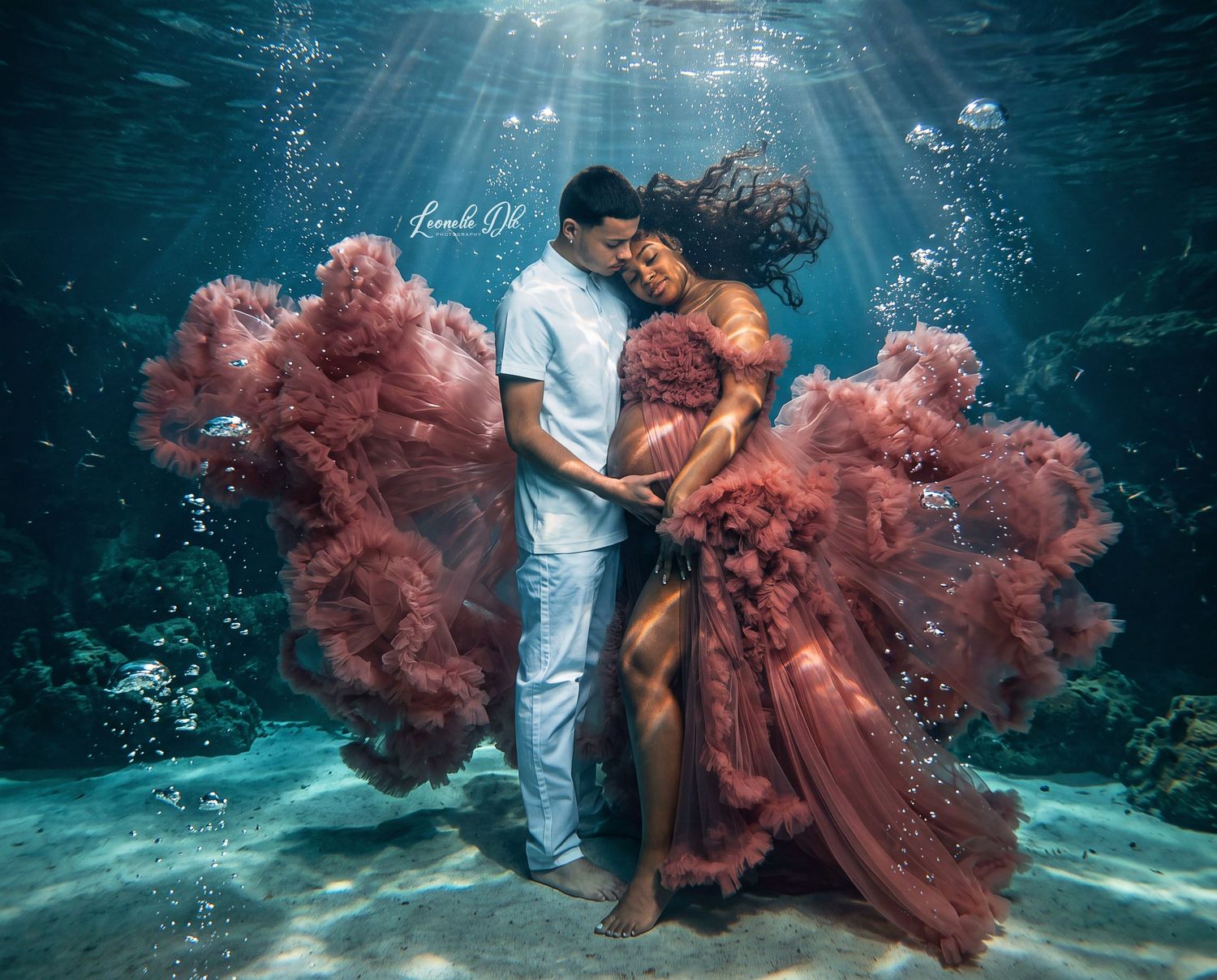 Couple embracing underwater in a stunning maternity photo shoot, woman wearing a flowing dusty rose gown with bubbles rising above.