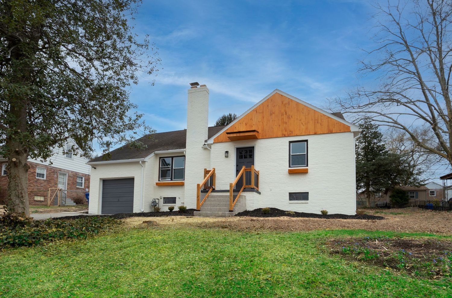 White brick ranch home with attached garage, wood deck, chimney, and green lawn surrounded by mature trees.
